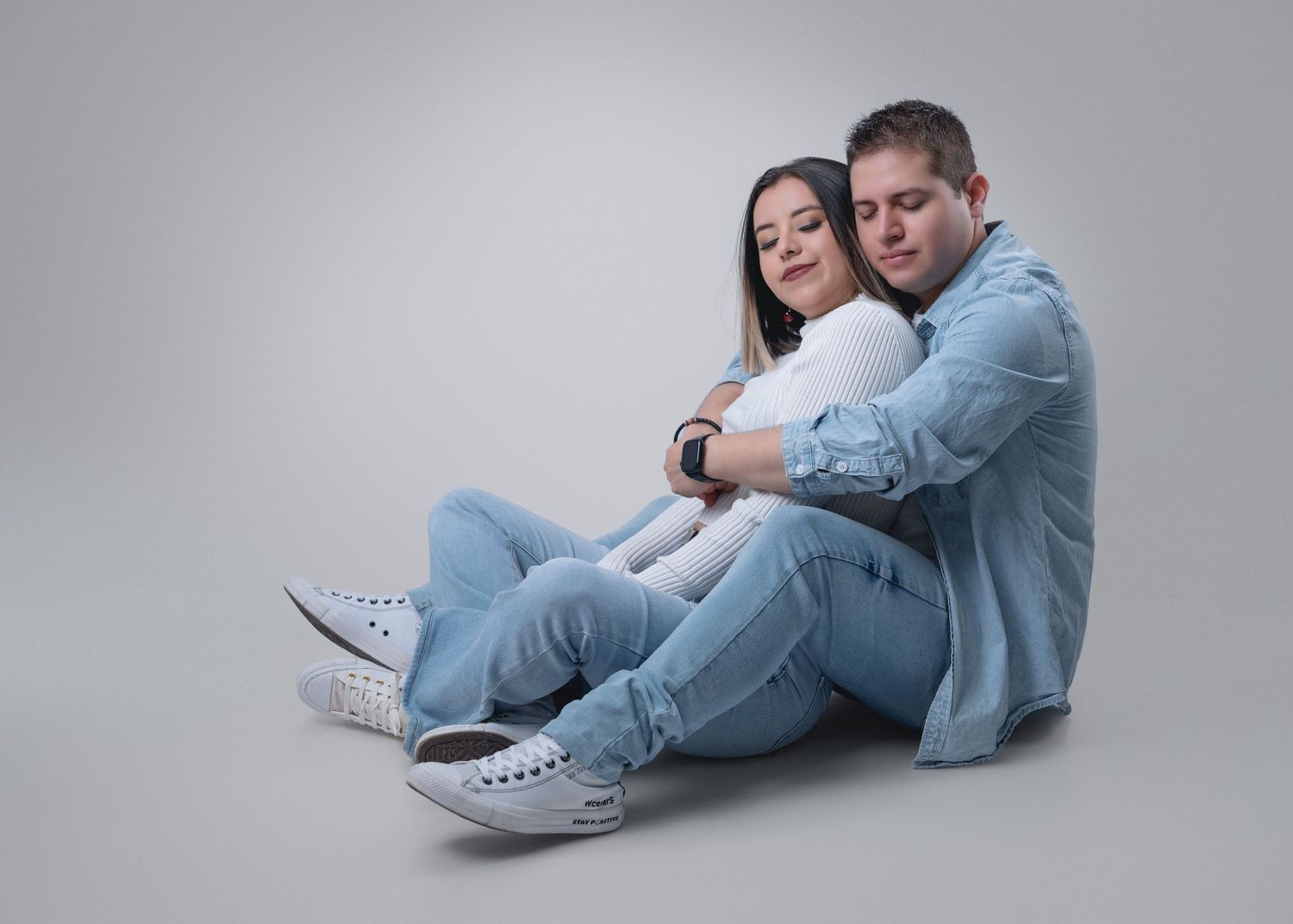 A couple in matching denim outfits and white sneakers sits together against a gray background in a studio setting.