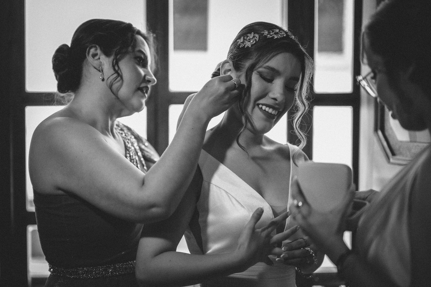 A black and white candid moment shows a bride and her friend sharing a joyful laugh during wedding preparations.