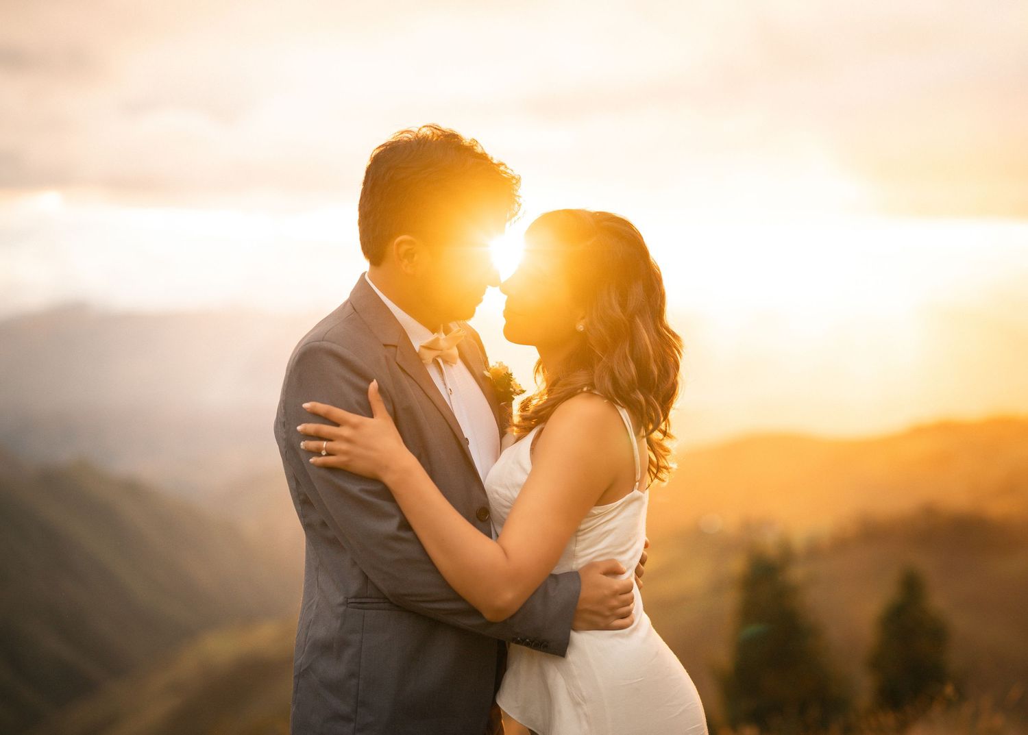 A couple embraces in a romantic moment at sunset with golden light streaming through mountains in the background.