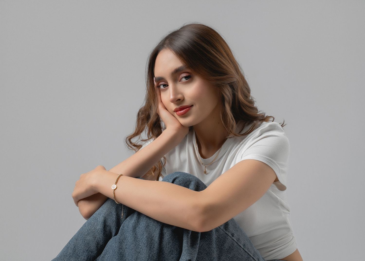 A person in a white t-shirt sits casually with arms resting on knees against a gray studio backdrop.