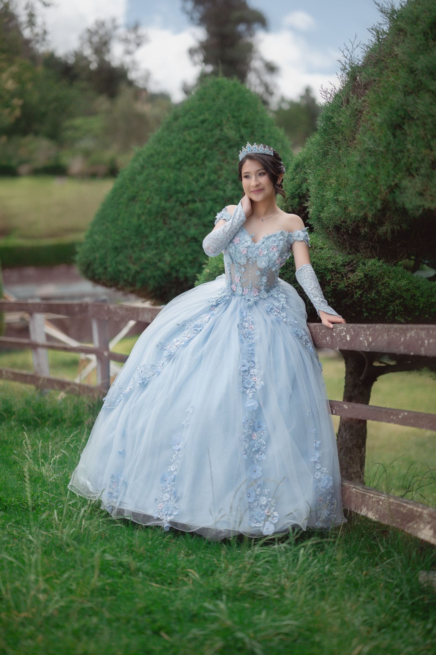 A dreamy light blue ball gown with floral details photographed in a lush garden setting with rustic wooden fencing.