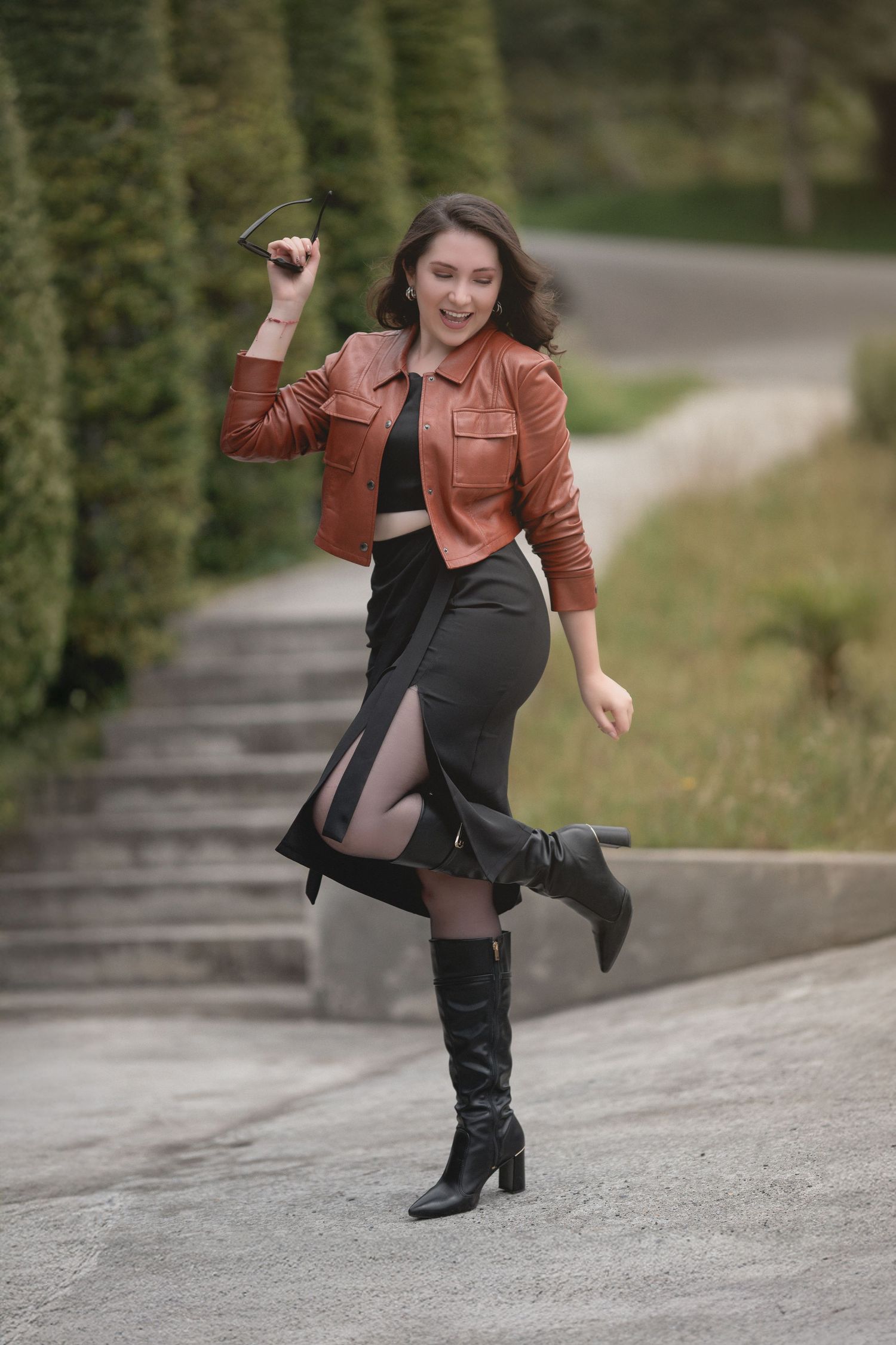 Stylish outfit featuring brown leather jacket, black skirt and knee-high boots captured in dynamic outdoor pose on path.