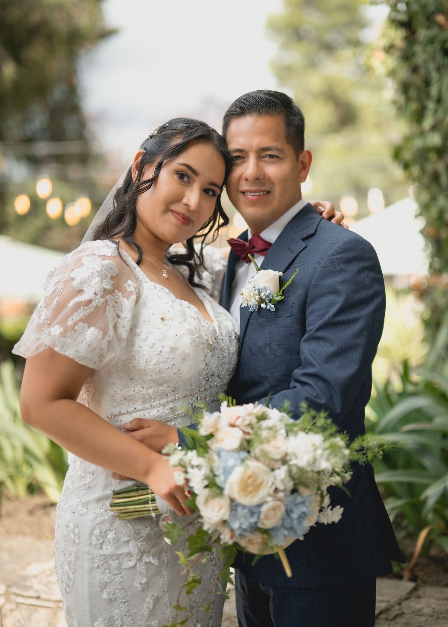 A romantic wedding couple embraces outdoors in a garden setting with soft natural lighting and floral bouquet details.