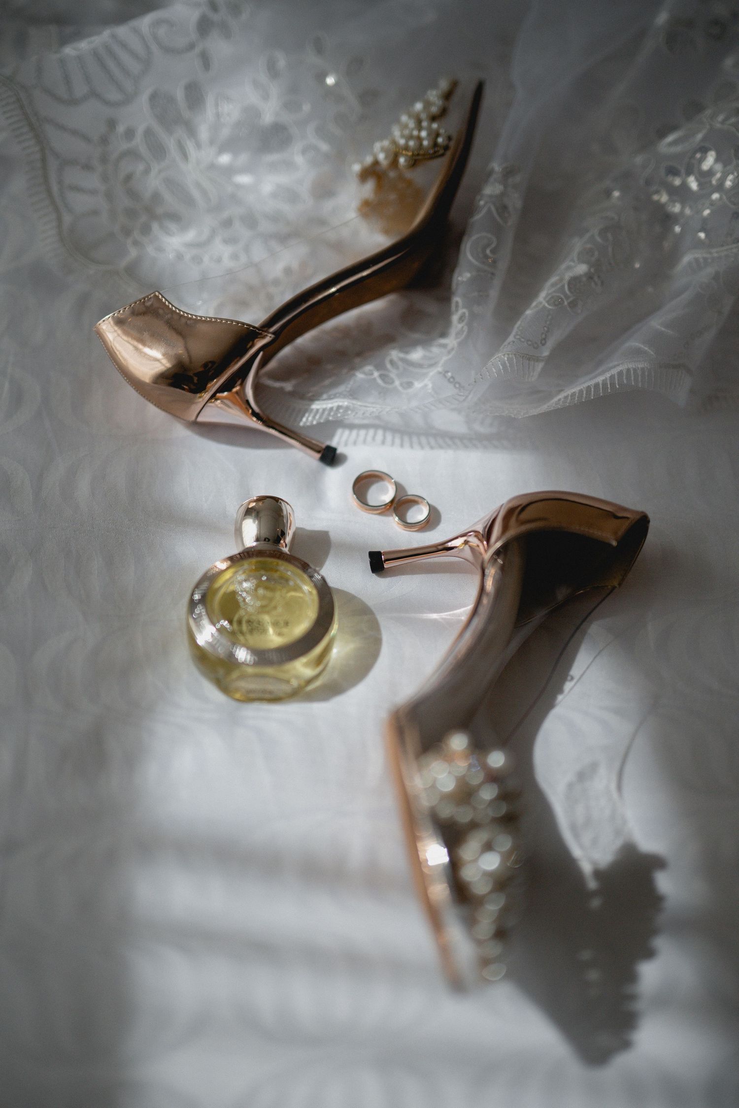 Wedding rings and perfume bottle arranged with delicate lace and a blush pink high heel shoe in soft lighting.