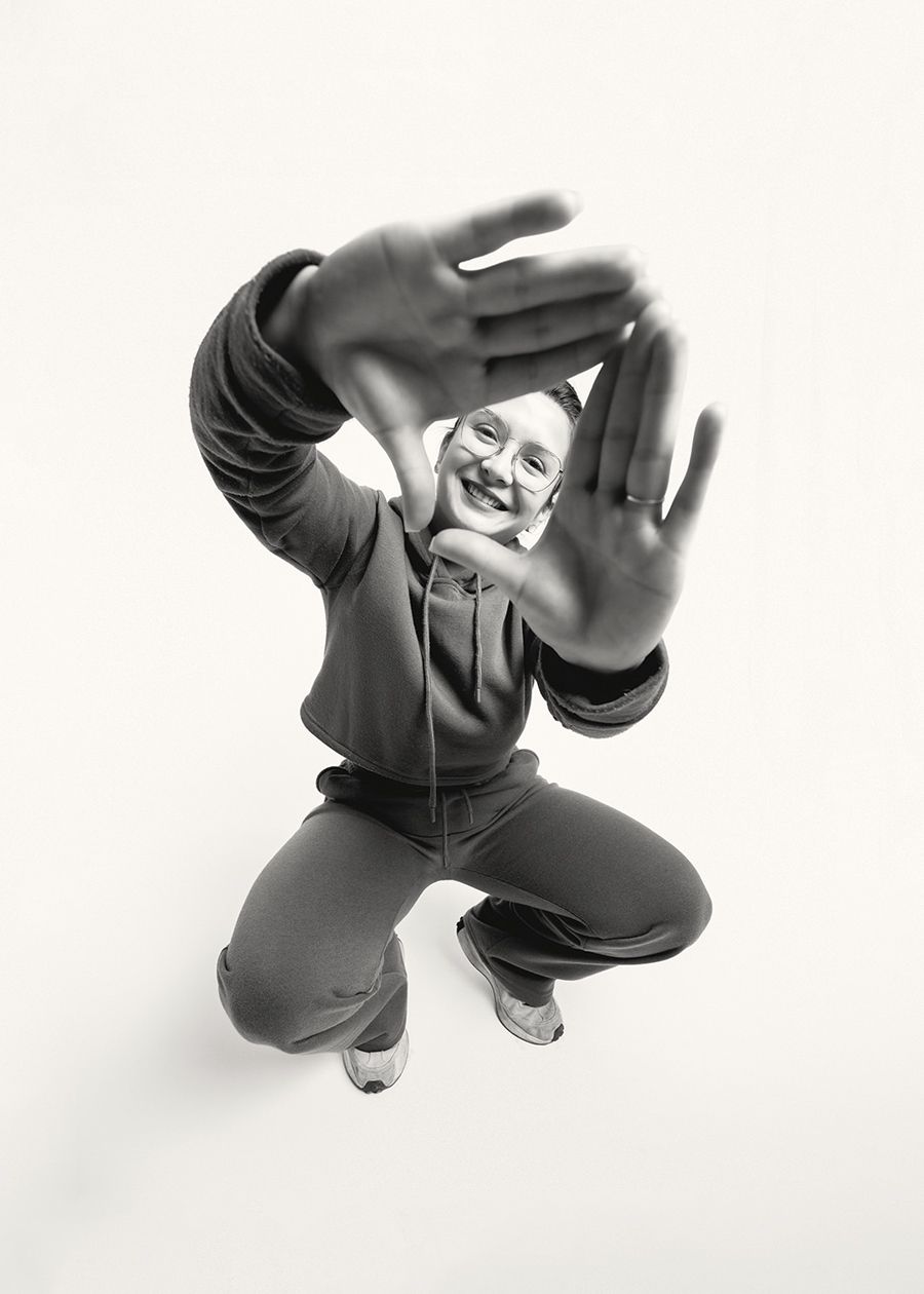 A person in a dark hoodie and pants crouches playfully with hands raised against a white background.