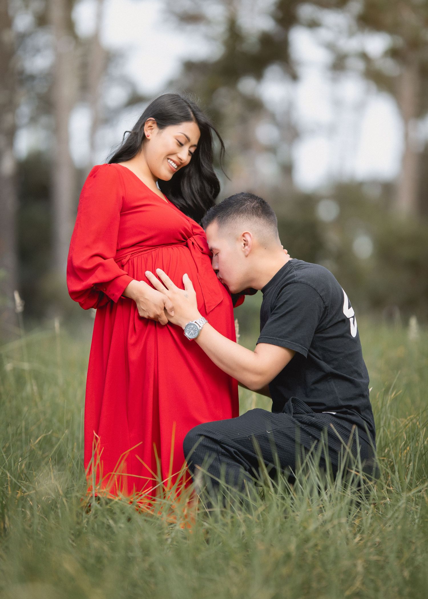 A couple in a grassy field shares an intimate moment as they pose for maternity photos in a red dress and black shirt.