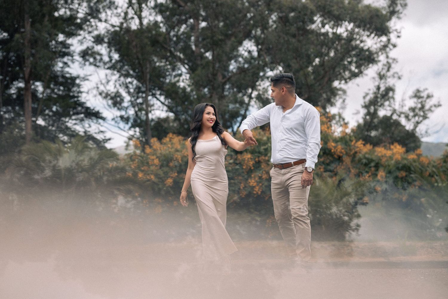A couple walks hand in hand through misty scenery with trees and mountains in the background.