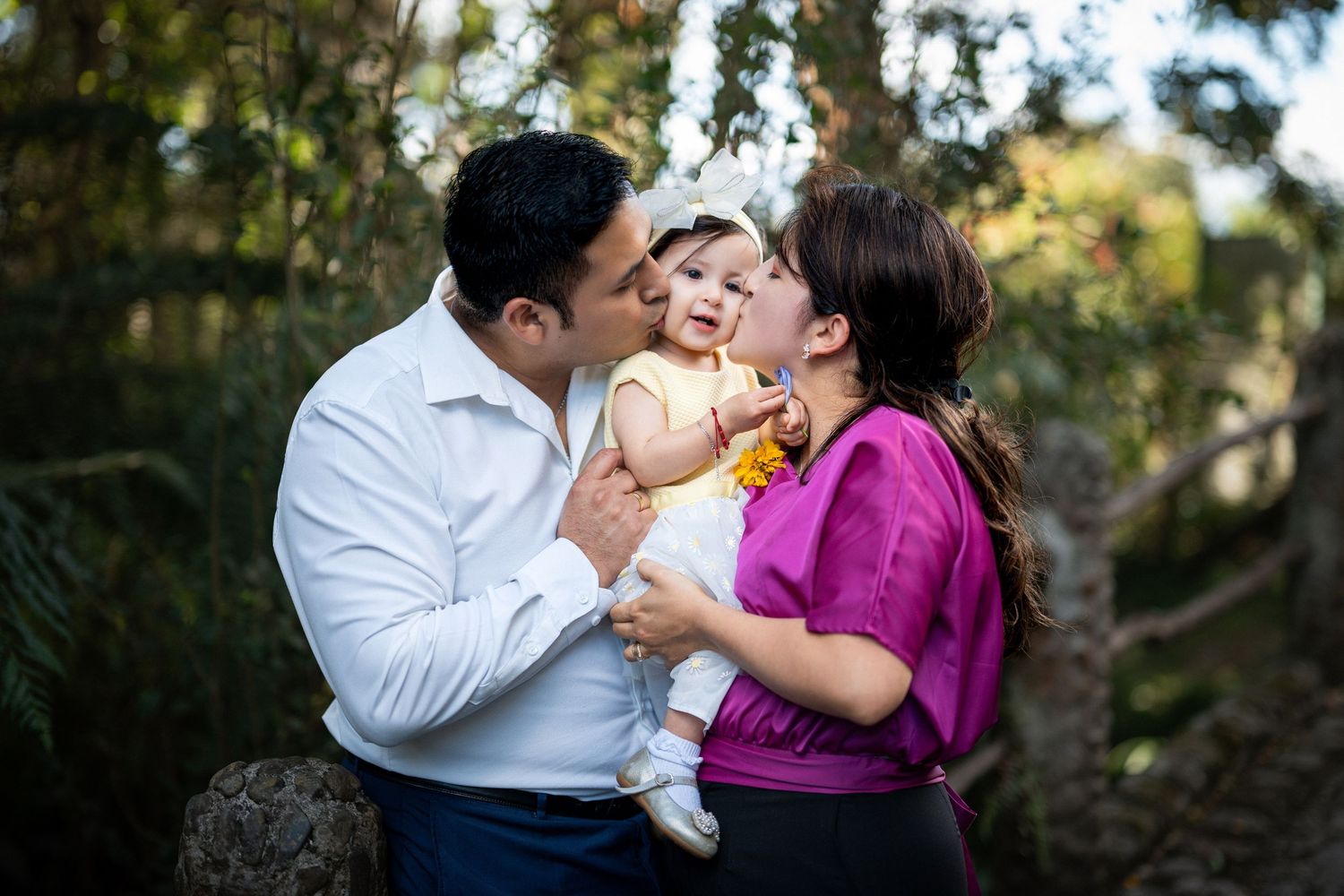 A couple embraces and kisses while holding a baby in a wooded outdoor setting during a family photo session.