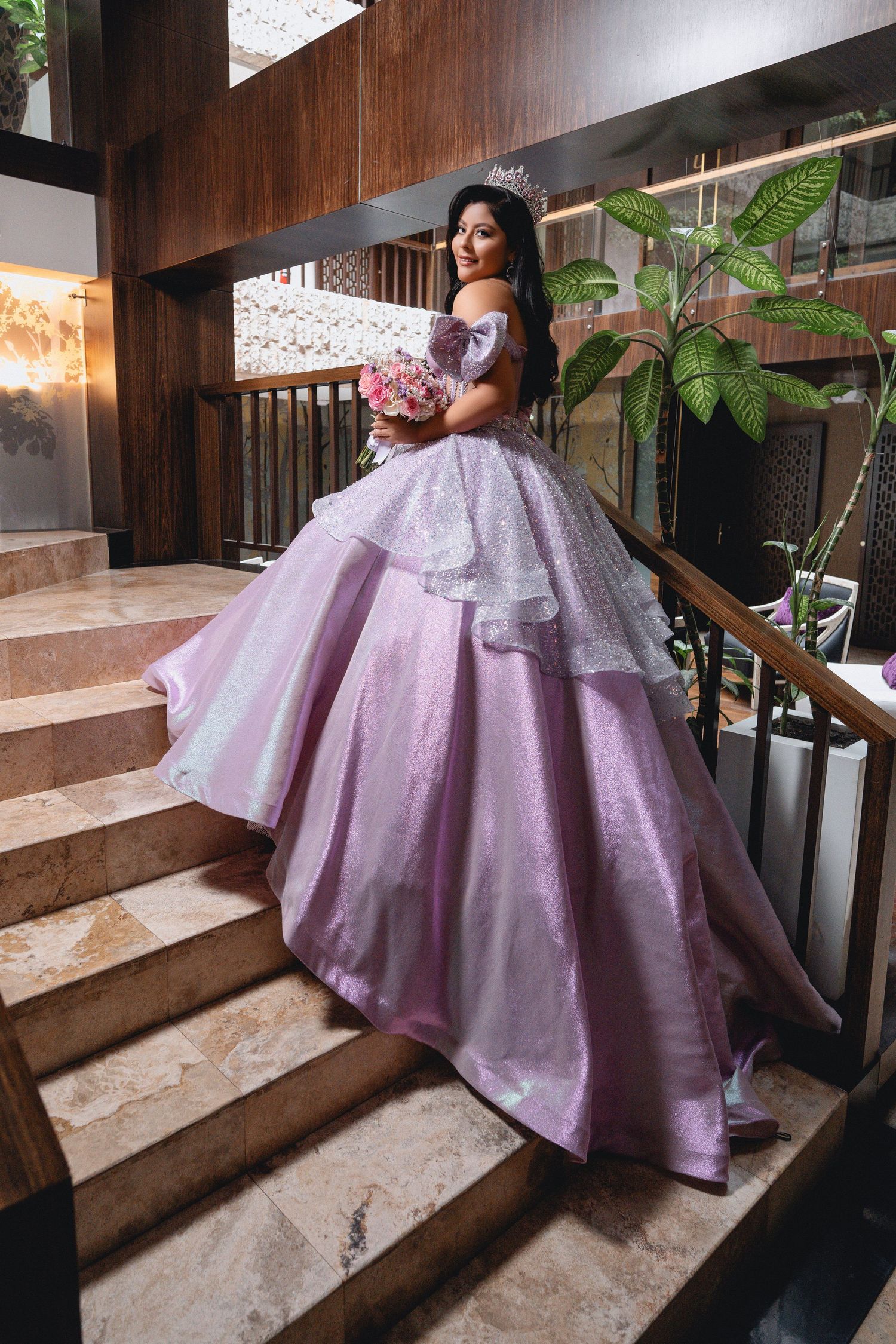 A stunning lavender ball gown with off-shoulder design and layered skirt photographed on marble stairs with green plants.