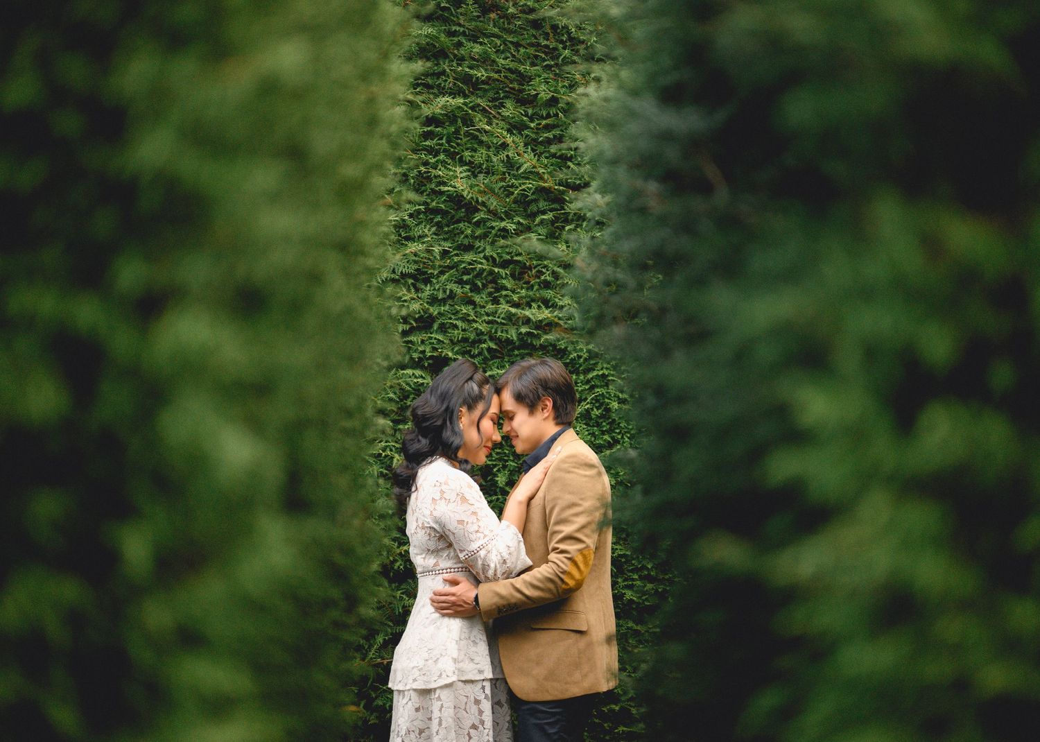 A romantic couple shares an intimate moment in front of lush green foliage in a garden setting.