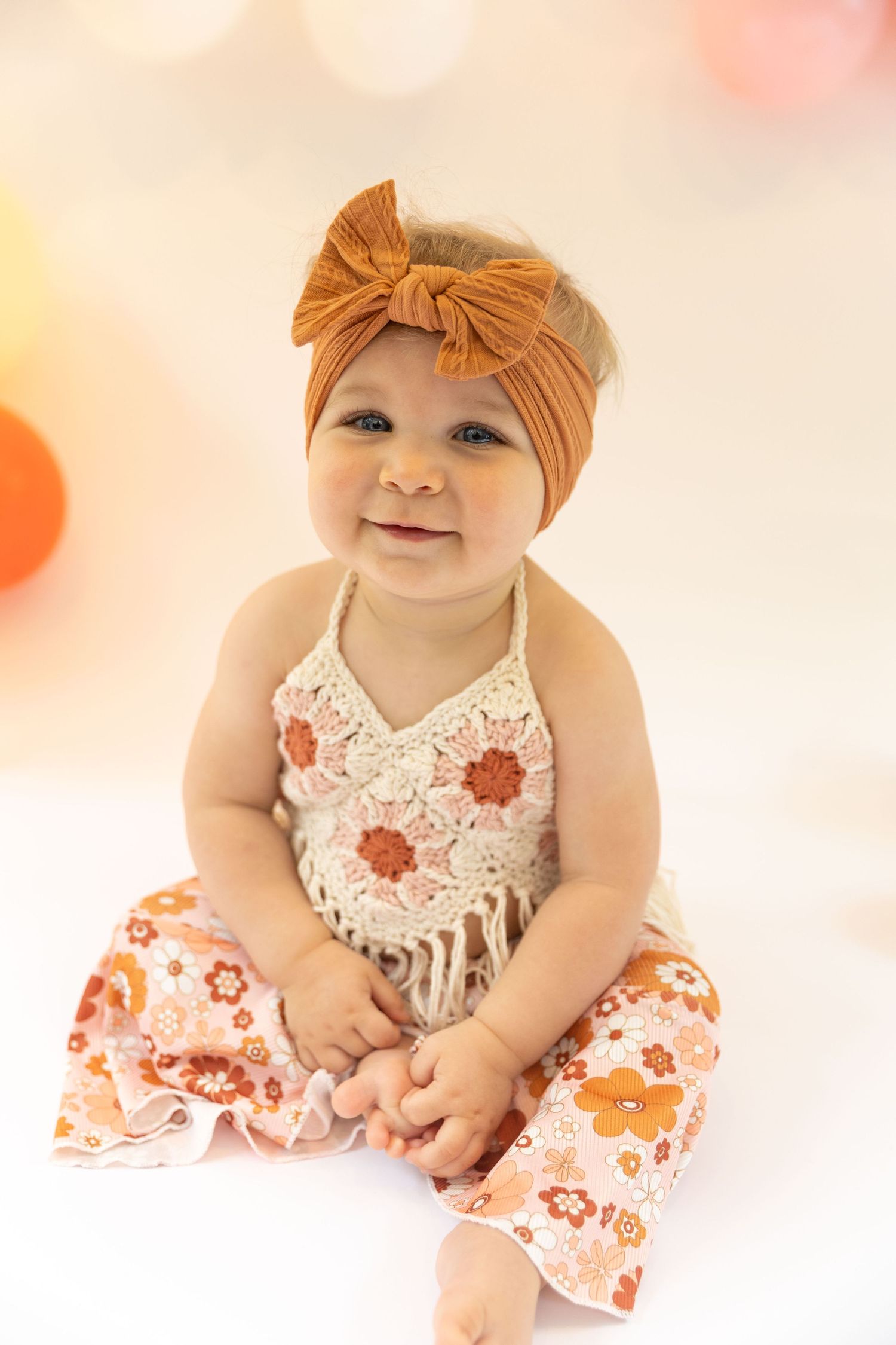 Baby wearing an orange bow headband and floral outfit smiles at the camera.