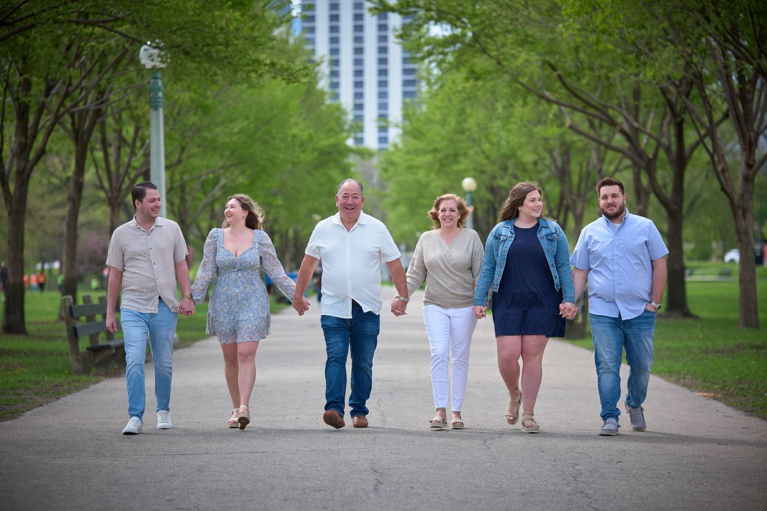 Six family members walk hand-in-hand down a tree-lined path in a sunny park, smiling together.