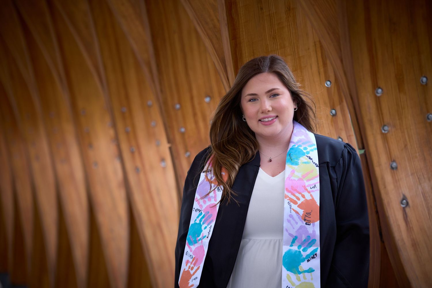 Graduate in black gown and colorful stole smiles confidently in front of a curved wooden wall.