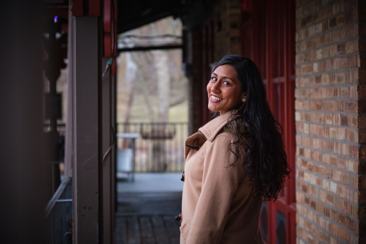Smiling woman in beige coat standing by red brick building, looking over shoulder outdoors.