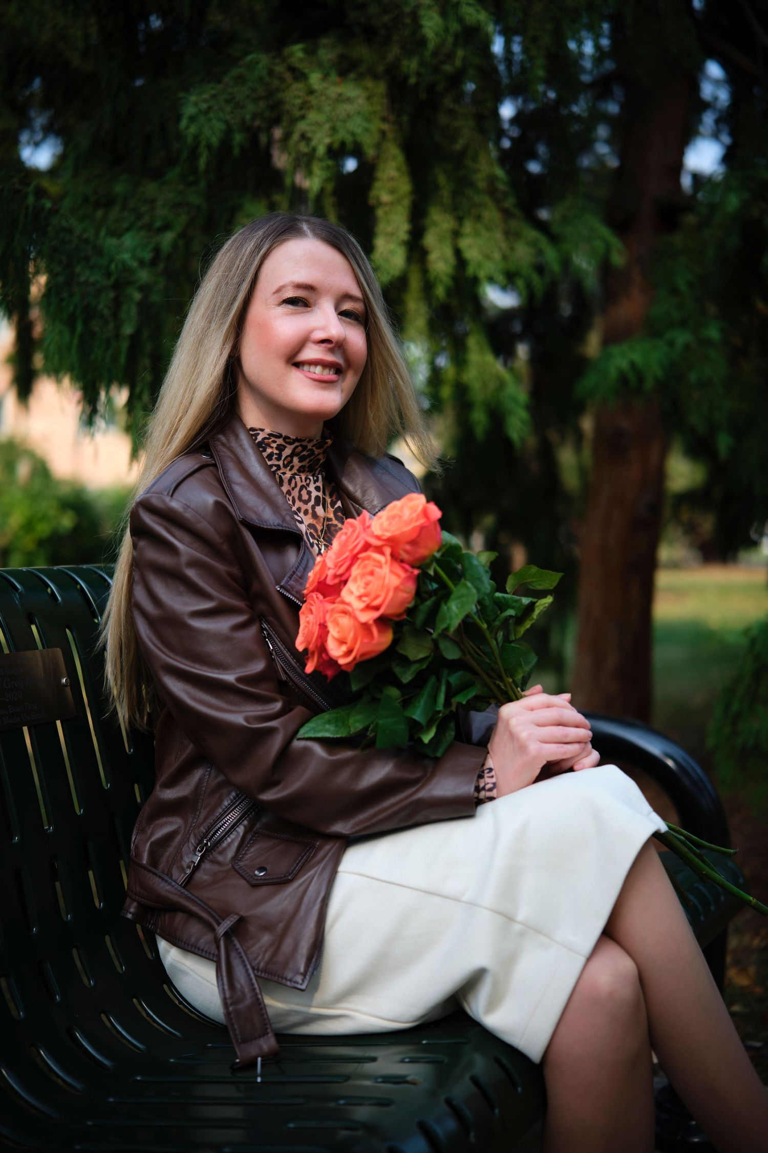 Smiling blonde woman in brown leather jacket holding orange roses while sitting on a park bench outdoors.