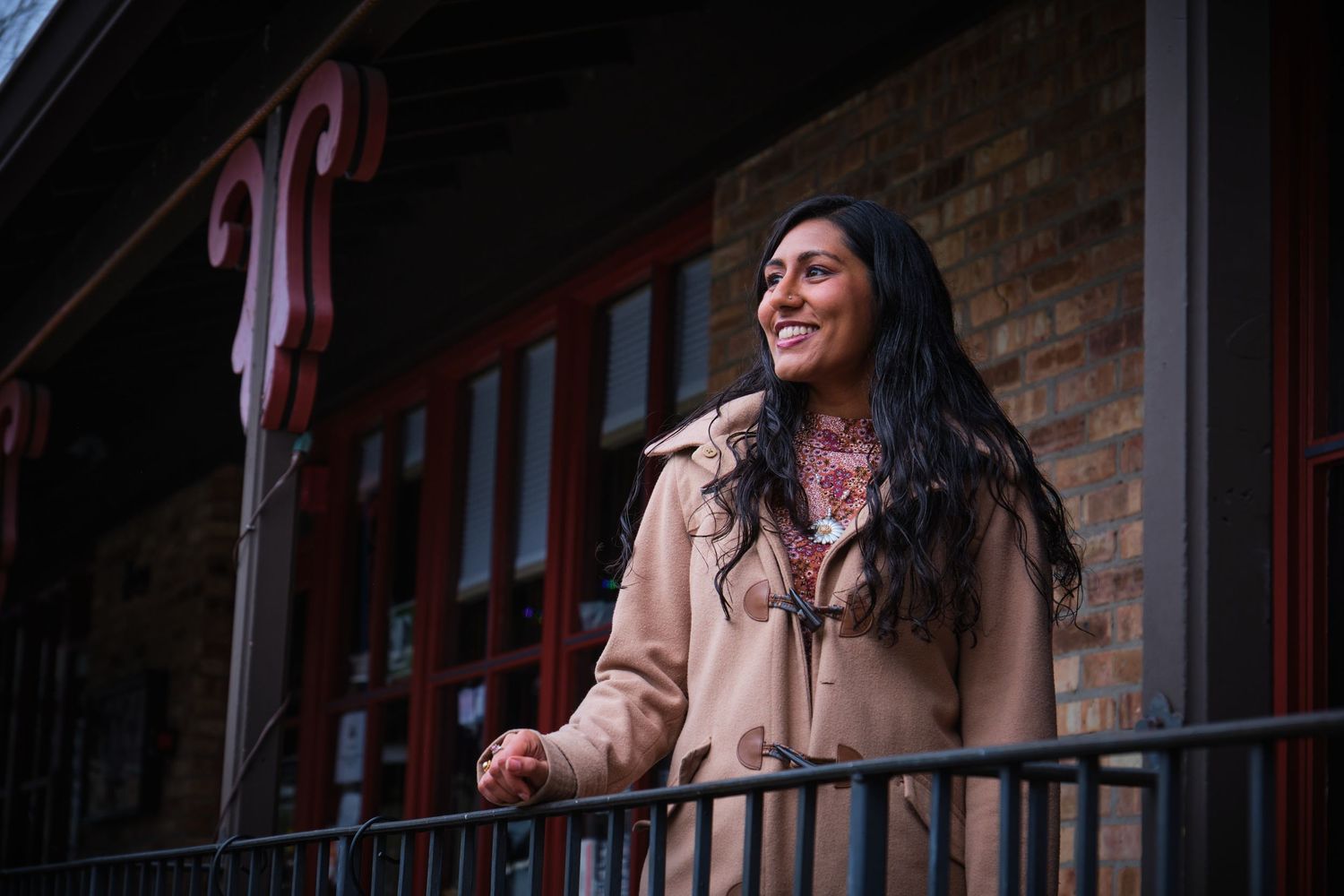 A smiling woman in a beige coat stands on a balcony outside a brick building with red decorative trim.