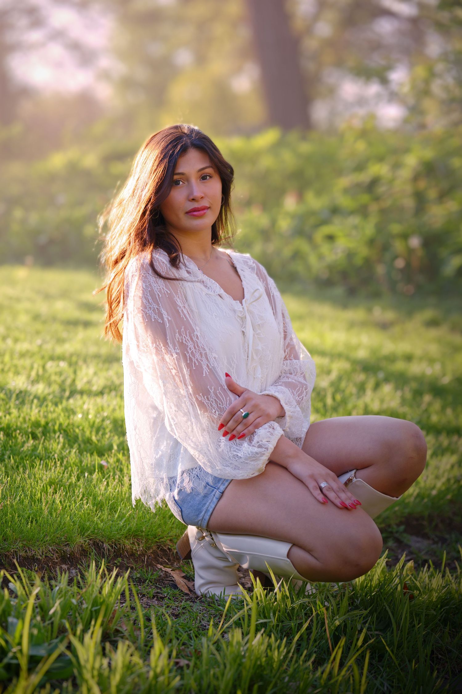 Young woman in white lace blouse and denim shorts kneeling on green grass in sunlit park setting.