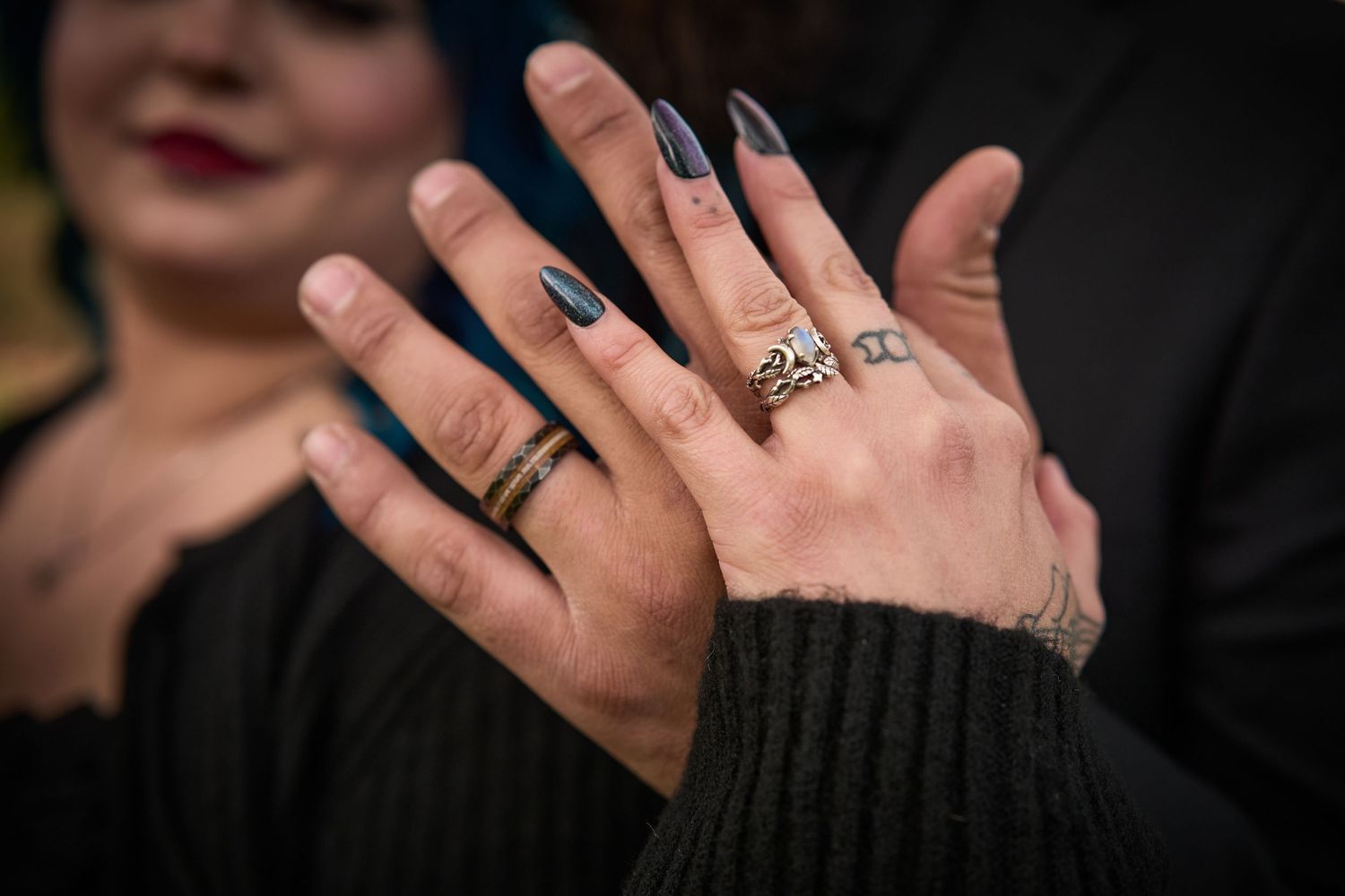 Close-up of hands displaying dark nail polish and engagement rings against black clothing.