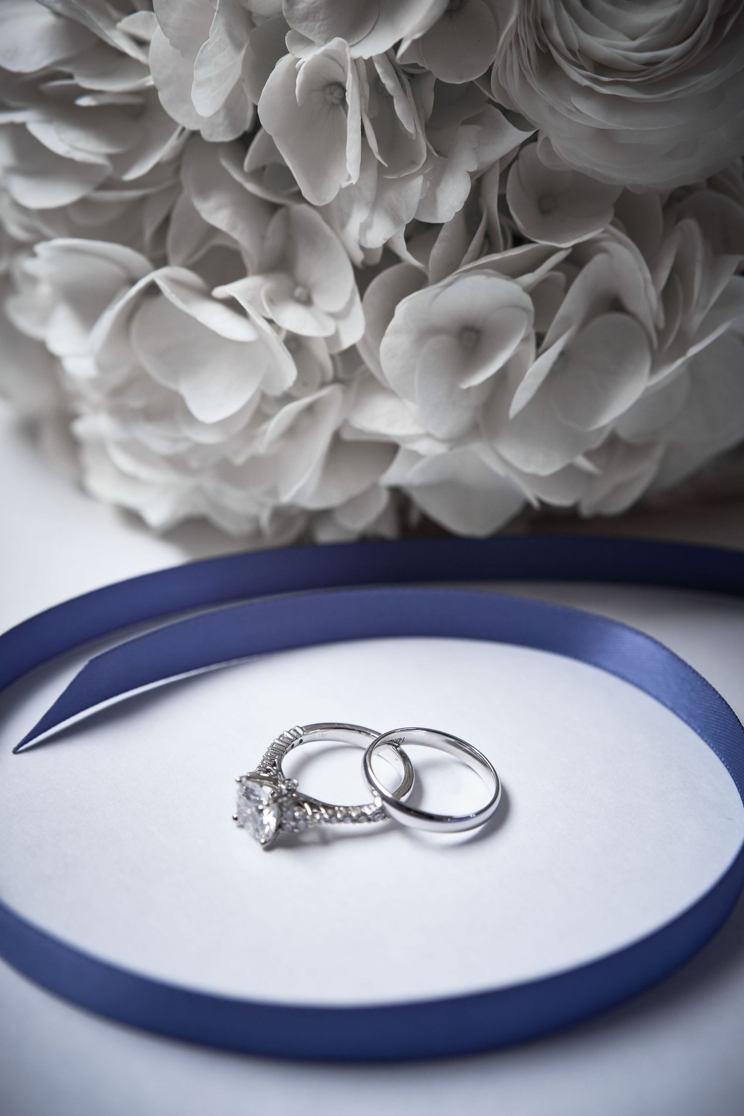 Wedding rings photographed on a navy blue and white surface with white flowers in the background.