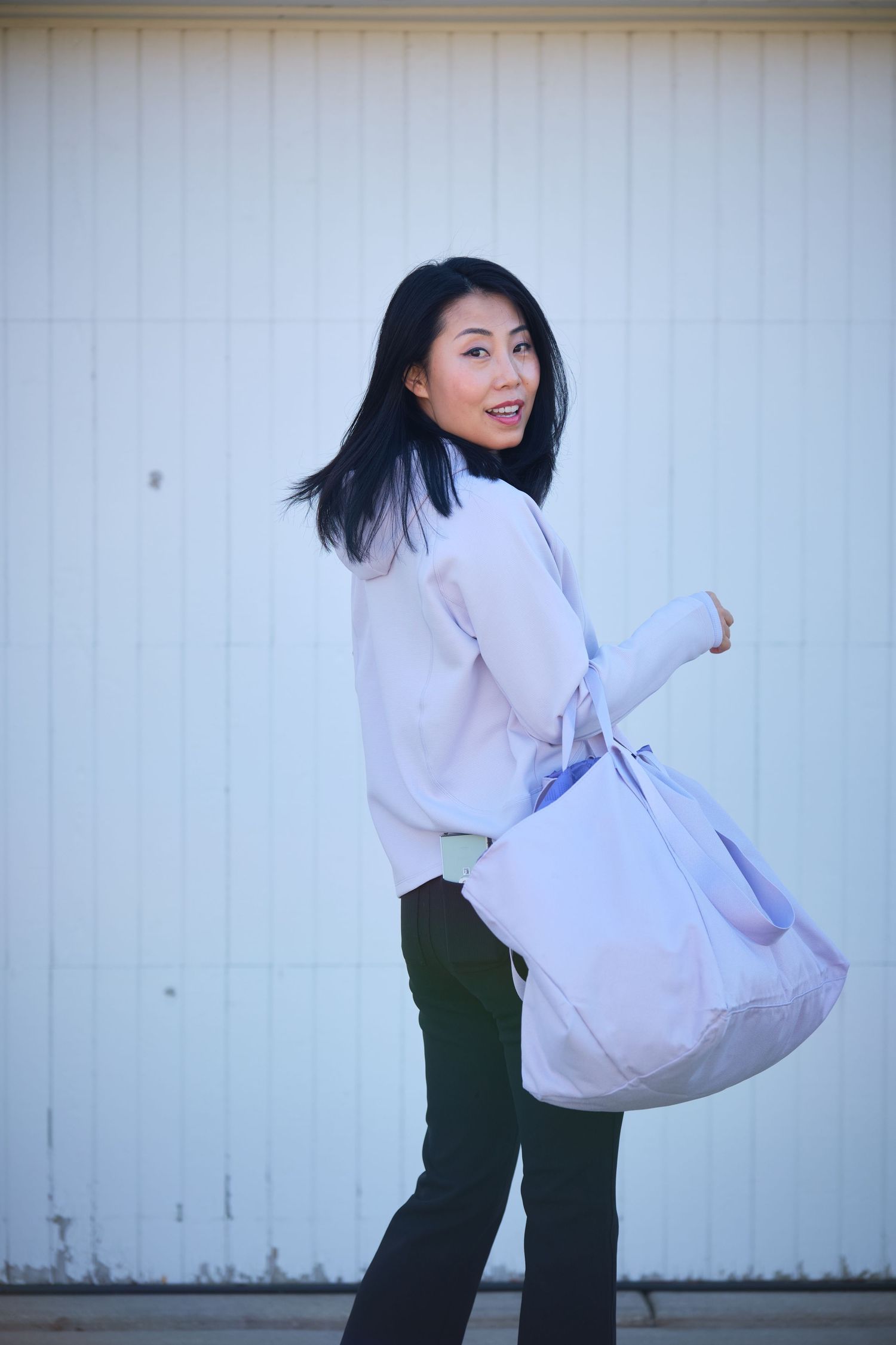 Asian woman in white sweater and dark pants carries a large lavender tote bag, smiling over her shoulder.