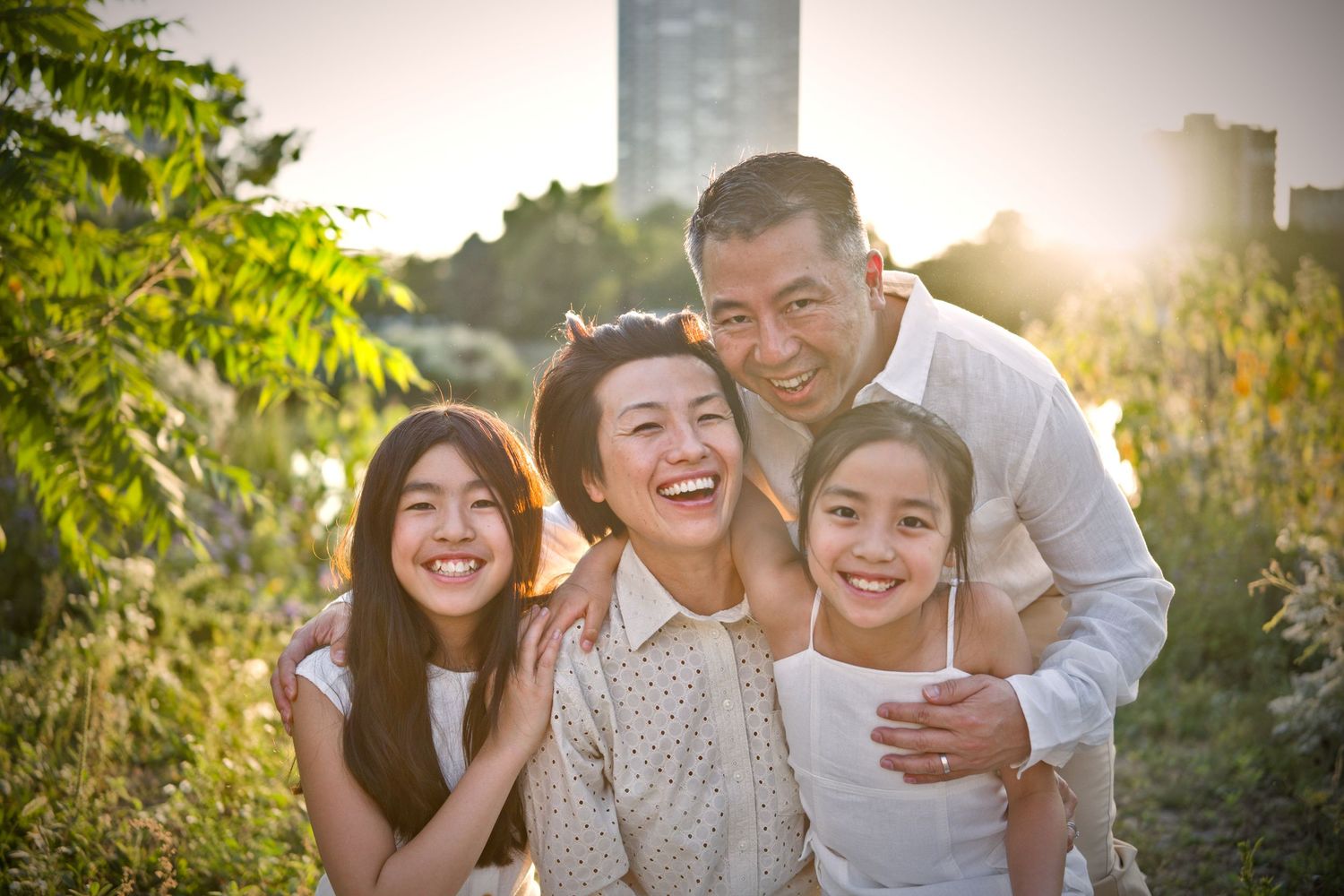 Happy Asian family of four smiling outdoors in golden sunlight, parents and two daughters posing together in a lush green park.