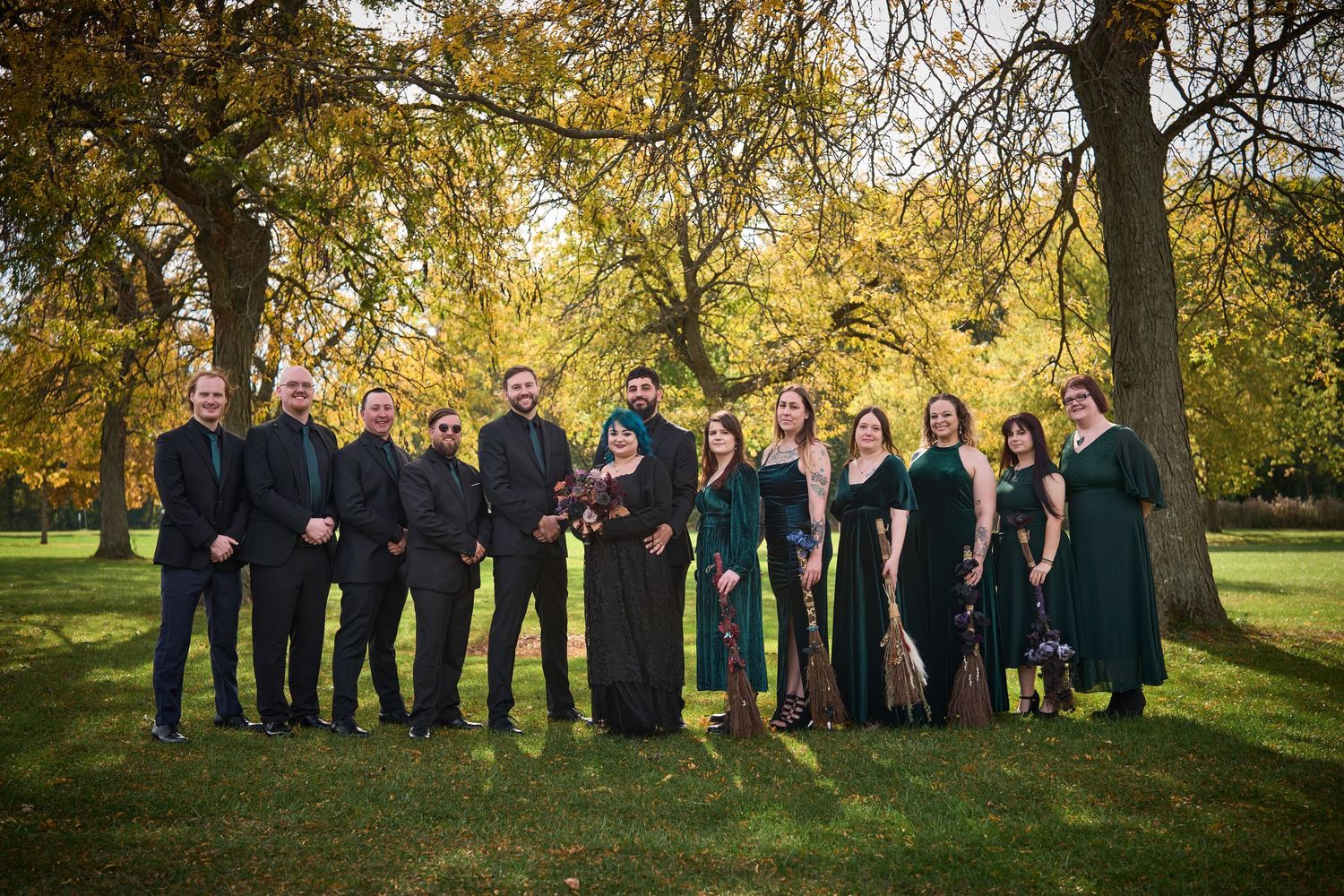 Large wedding party in formal black attire poses together under autumn trees in natural lighting.