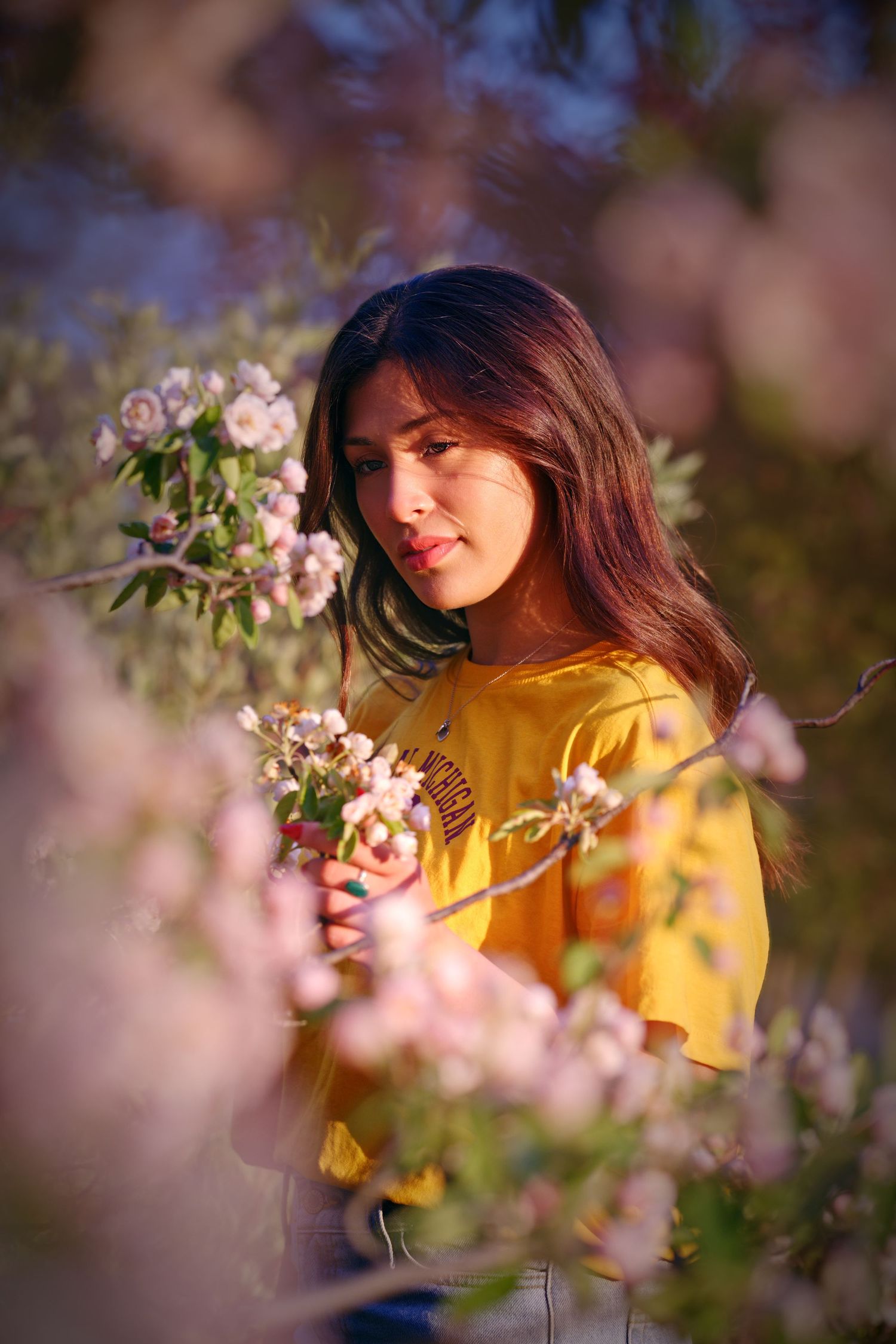 Young woman in yellow top surrounded by pink blossoms in golden hour light, looking down thoughtfully.