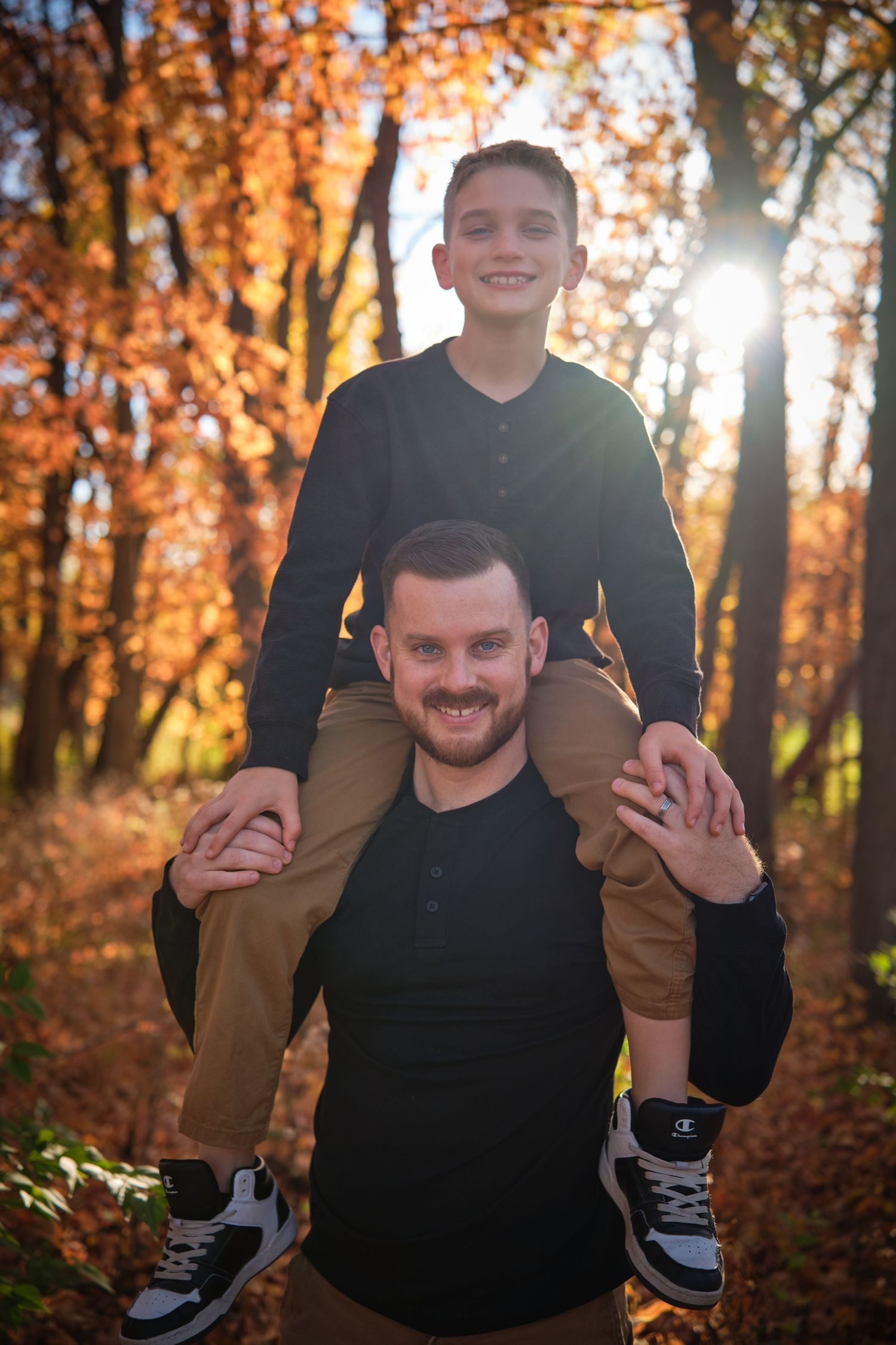 A smiling boy sits on a man's shoulders in a sunlit autumn forest, both wearing black tops and khaki pants.