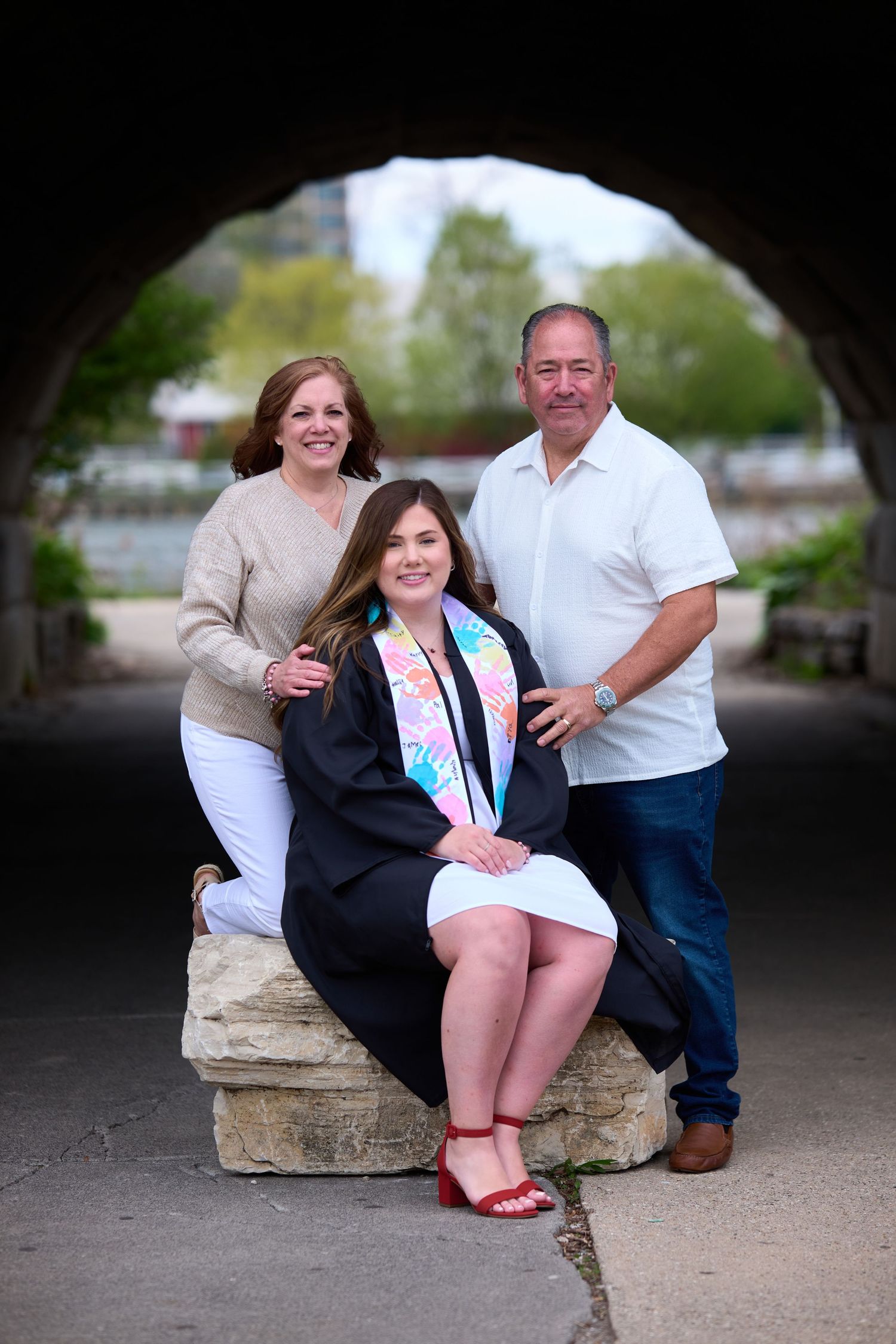 A graduate in cap and gown poses with two proud parents under a stone arch bridge outdoors.