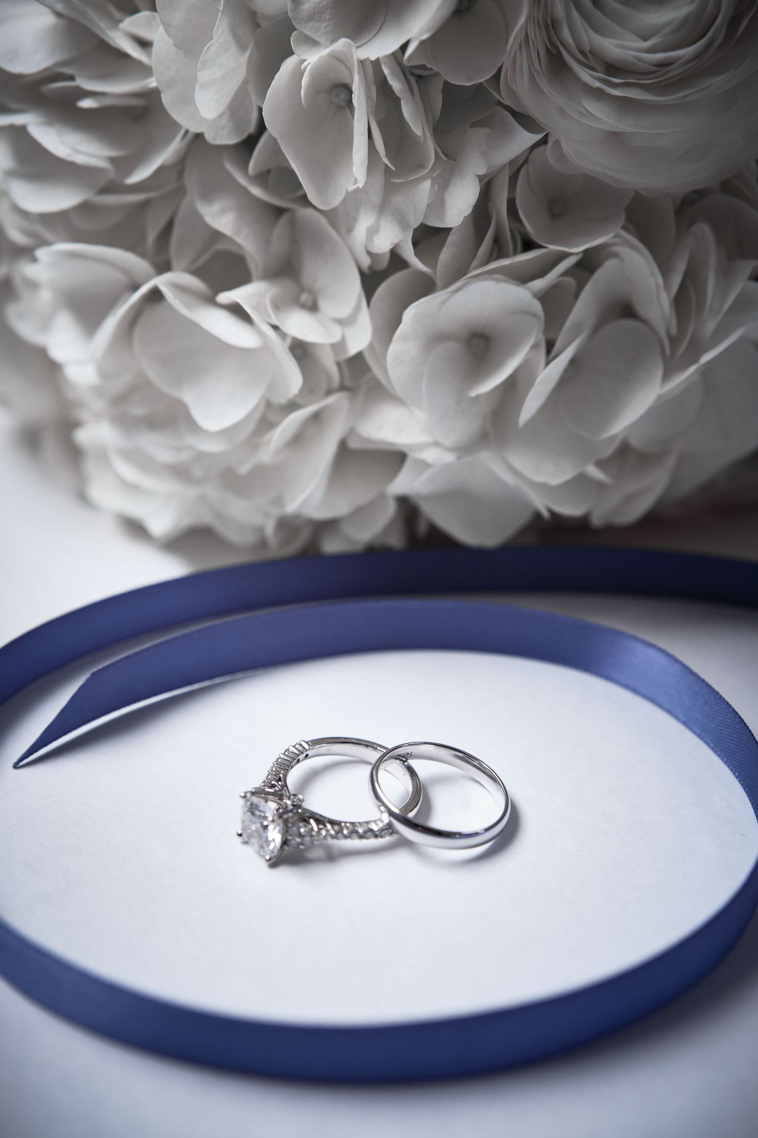 Wedding rings displayed on navy blue and white surface with soft floral backdrop.