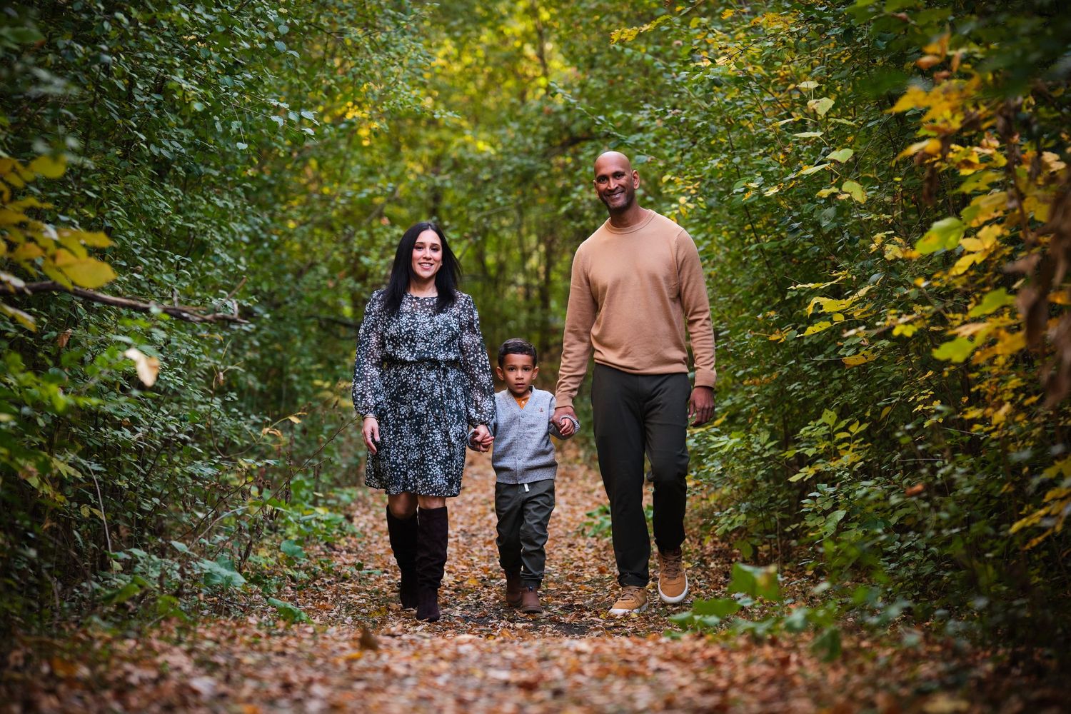 Family of three walking hand-in-hand on a leaf-covered autumn forest path surrounded by green and yellow foliage.