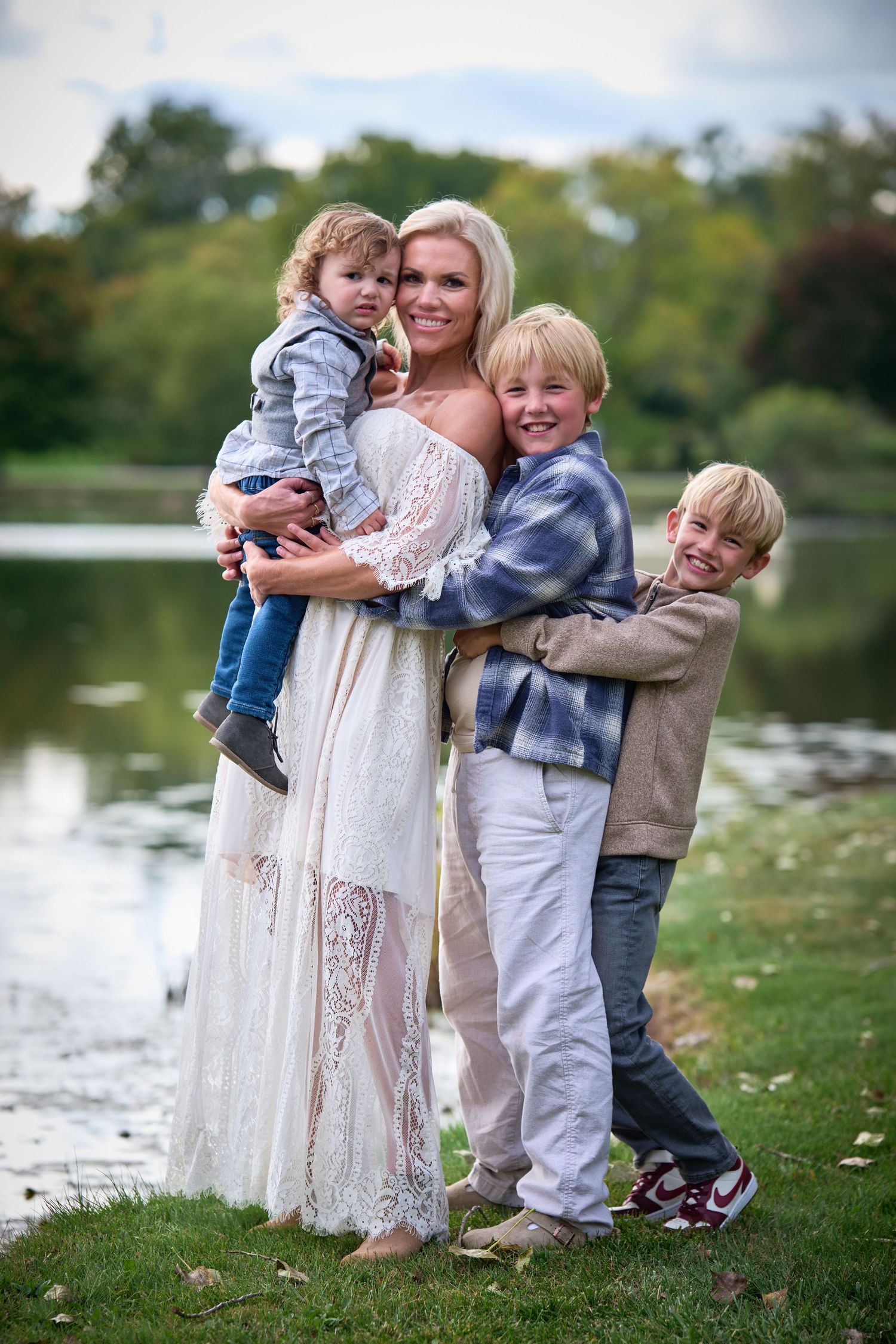 Blonde woman in white lace dress holds toddler while two boys hug her outdoors near a pond.