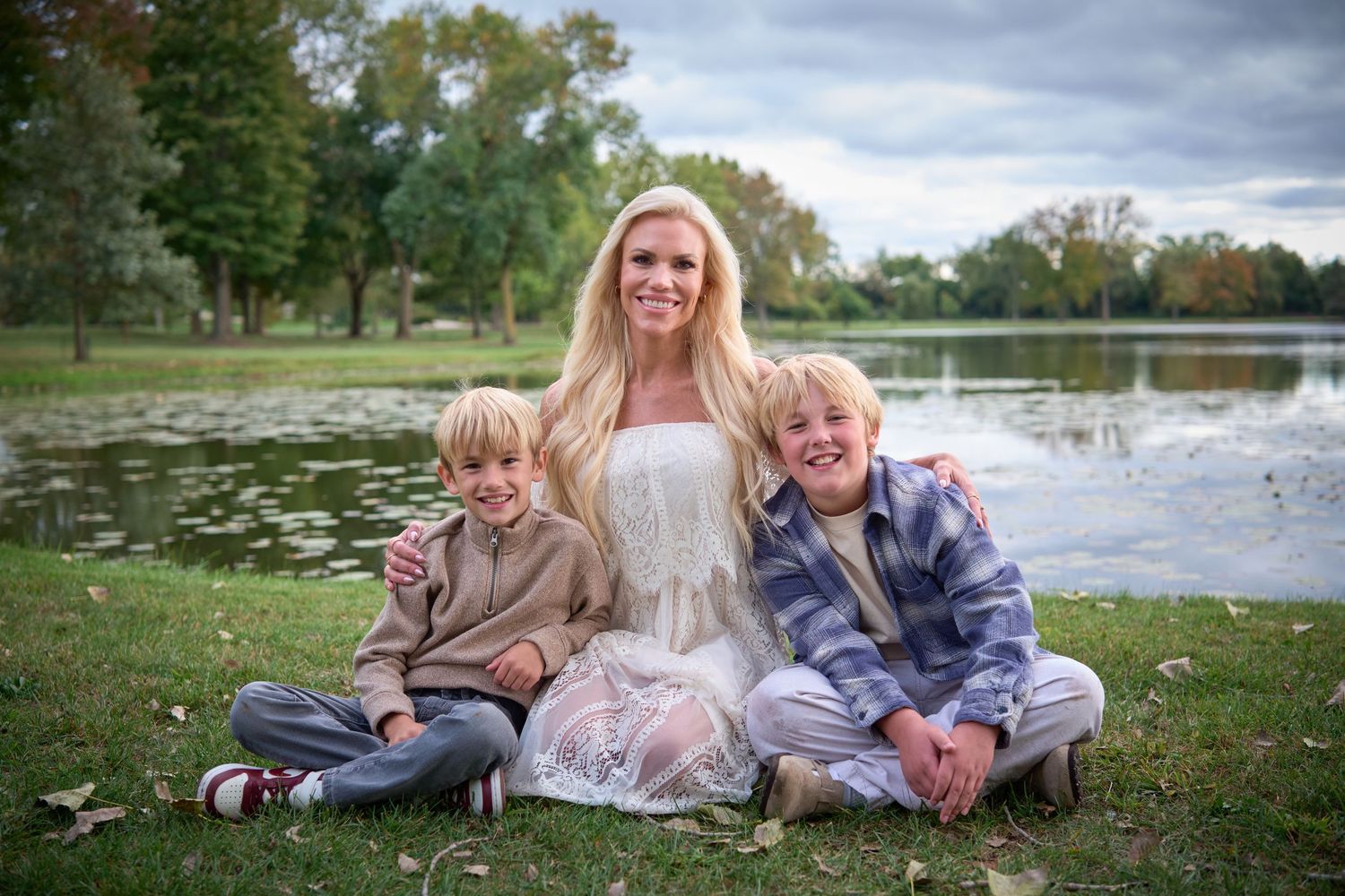 Smiling blonde woman sitting on grass with two young boys near a scenic pond in a park during fall.