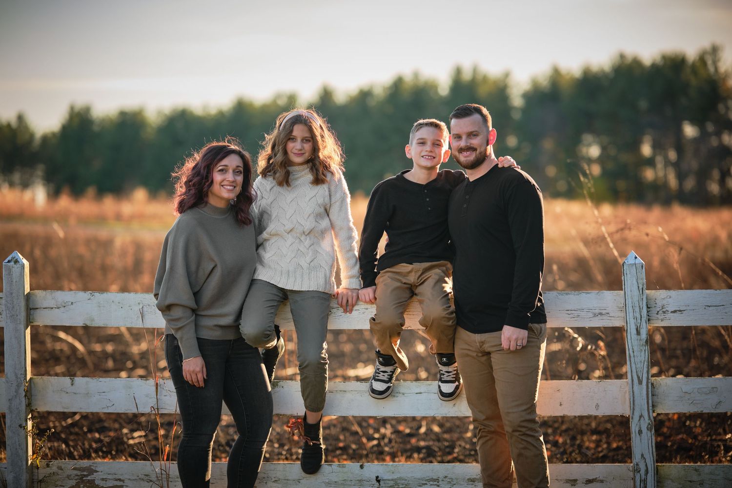 Family of four posing by a rustic wooden fence in a golden field at sunset.