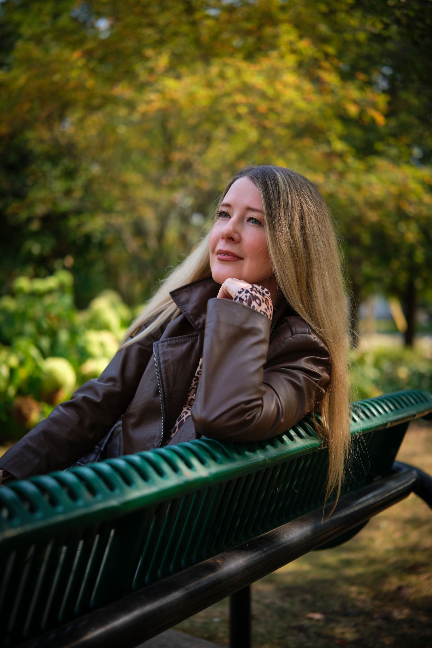 A woman with long blonde hair sits relaxed on a green park bench, wearing a brown leather jacket on an autumn day.