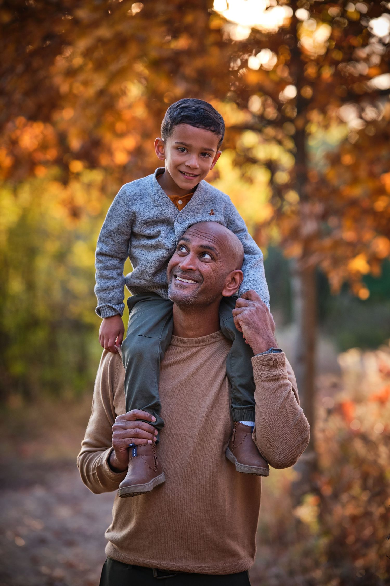 A smiling father carries his young son on his shoulders during an autumn walk among golden-orange trees.