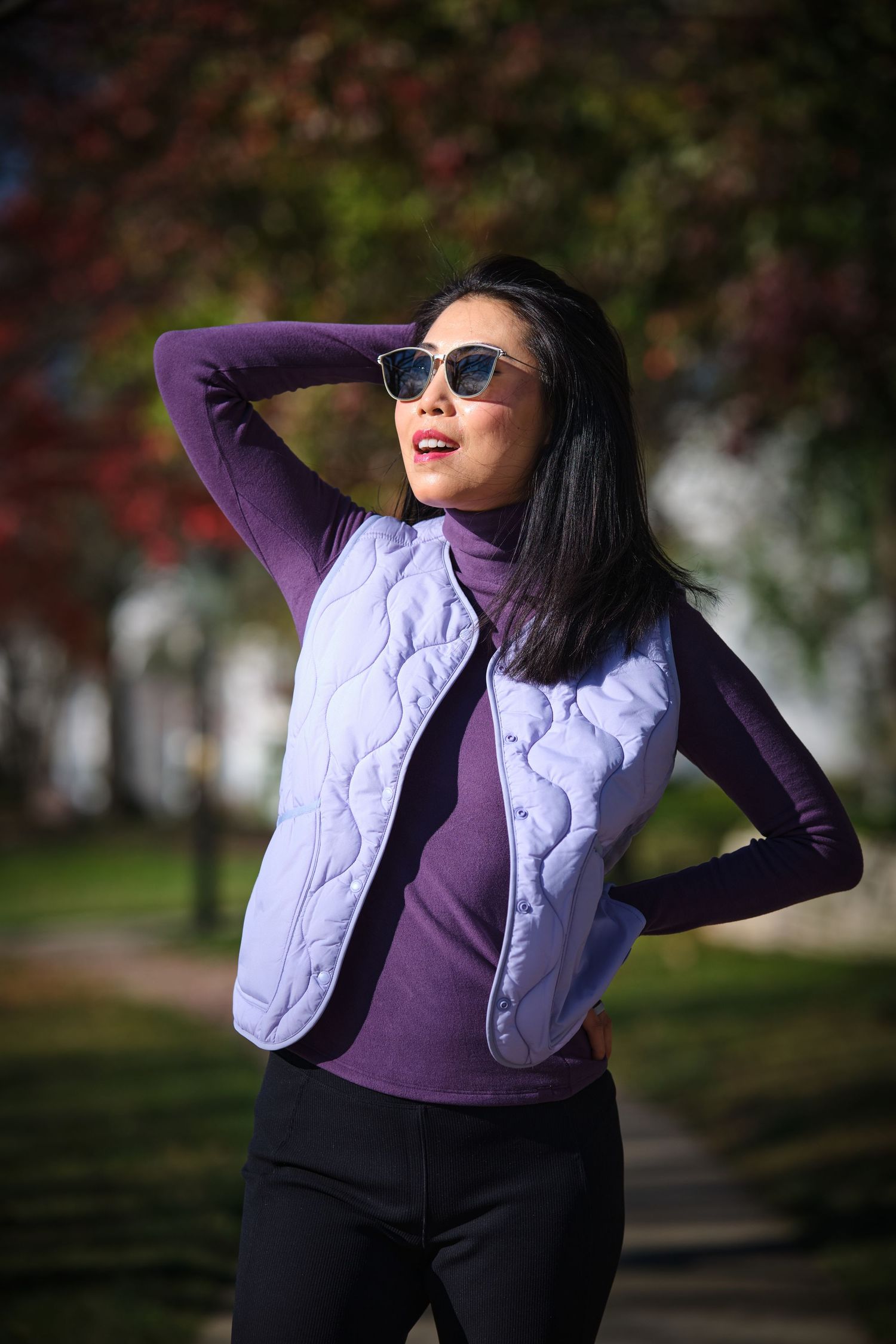 A stylish woman in a purple turtleneck and quilted vest poses outdoors wearing sunglasses in an autumn park.
