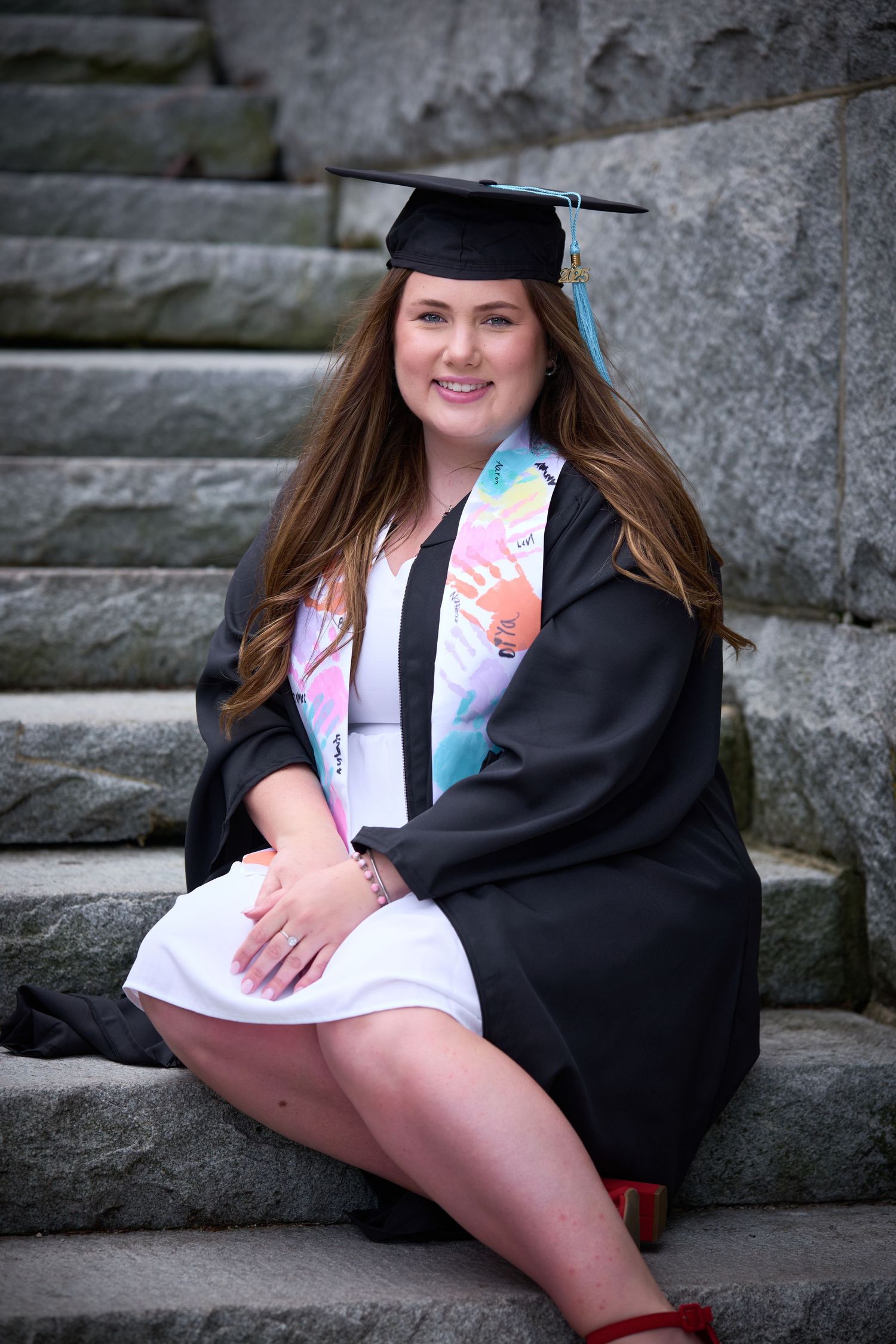 A smiling young woman in black graduation cap and gown with colorful stole sits on stone steps for graduation photos.