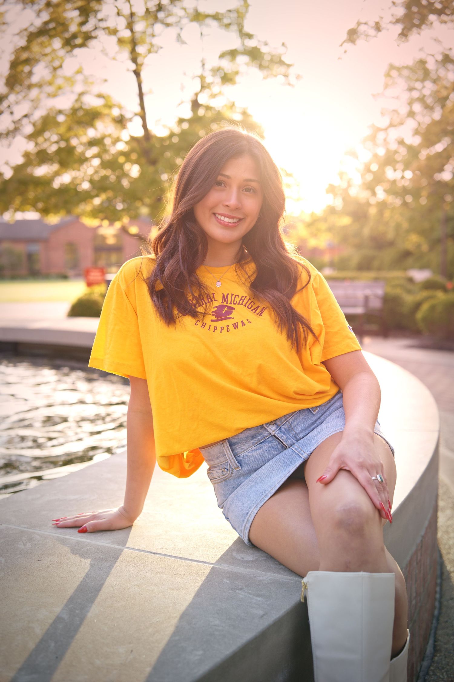 Young woman in Central Michigan Chippewas yellow t-shirt and denim skirt sitting outdoors near water at golden hour.