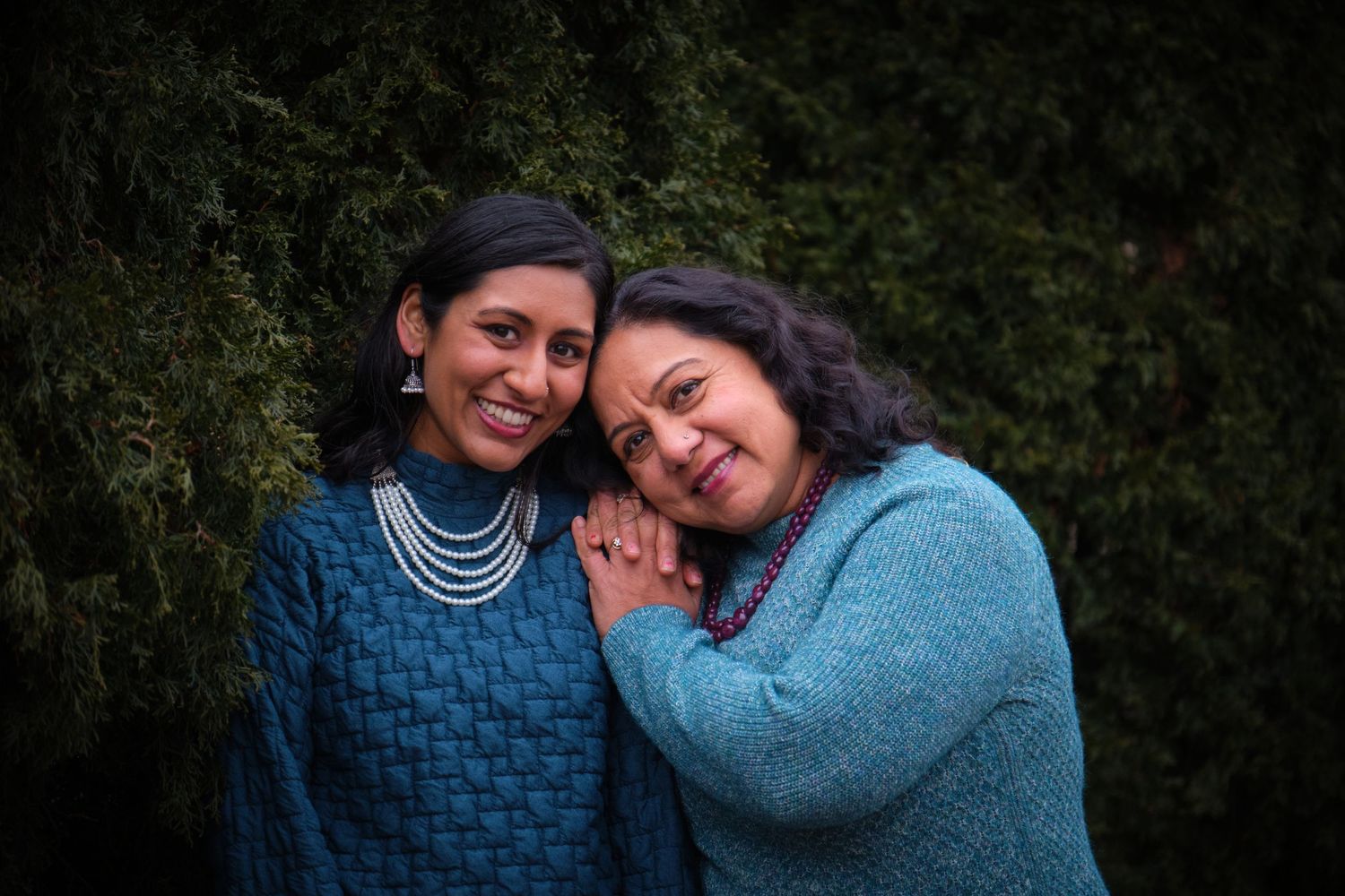 Two women in blue sweaters smiling and posing together outdoors with green trees in the background.