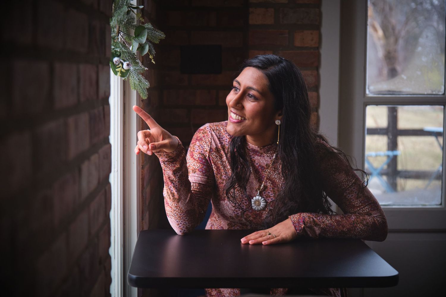 Smiling woman in floral dress sitting at dark wood table by brick wall window, pointing and looking outside.
