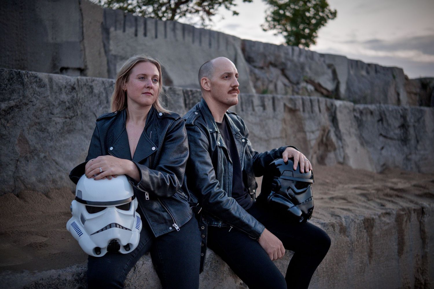 Two people in black leather jackets pose outdoors, one holding a Star Wars Stormtrooper helmet.