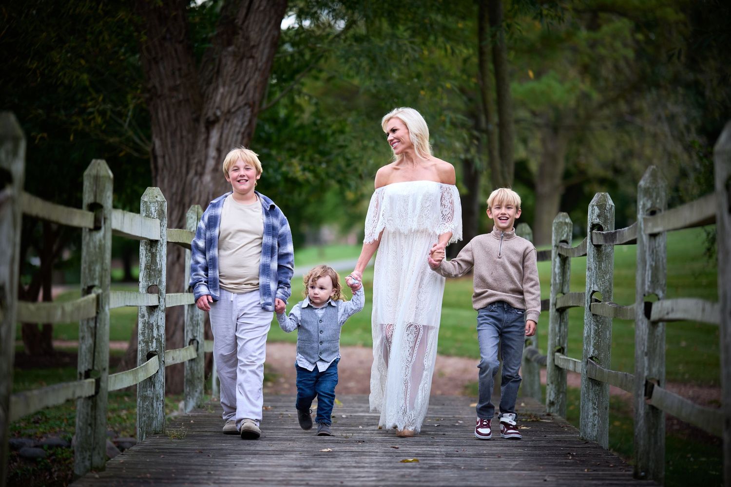 Family of four walking on a wooden bridge in a lush park, mother in white dress holding hands with children.
