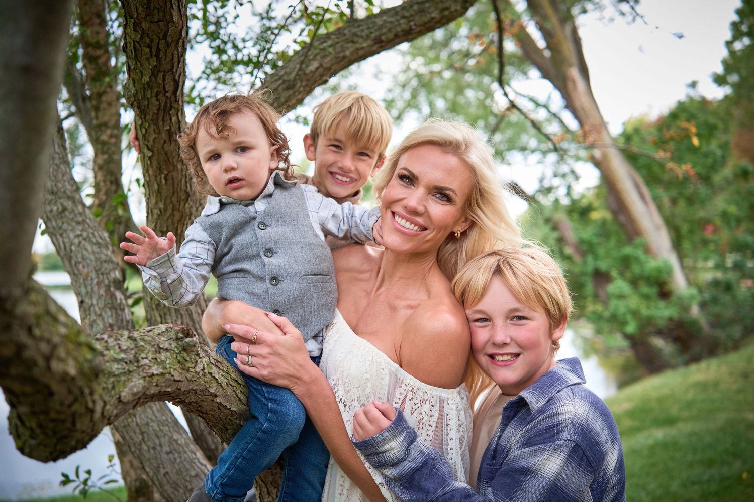 A smiling woman poses outdoors with three young boys near a tree in a lush green park setting.
