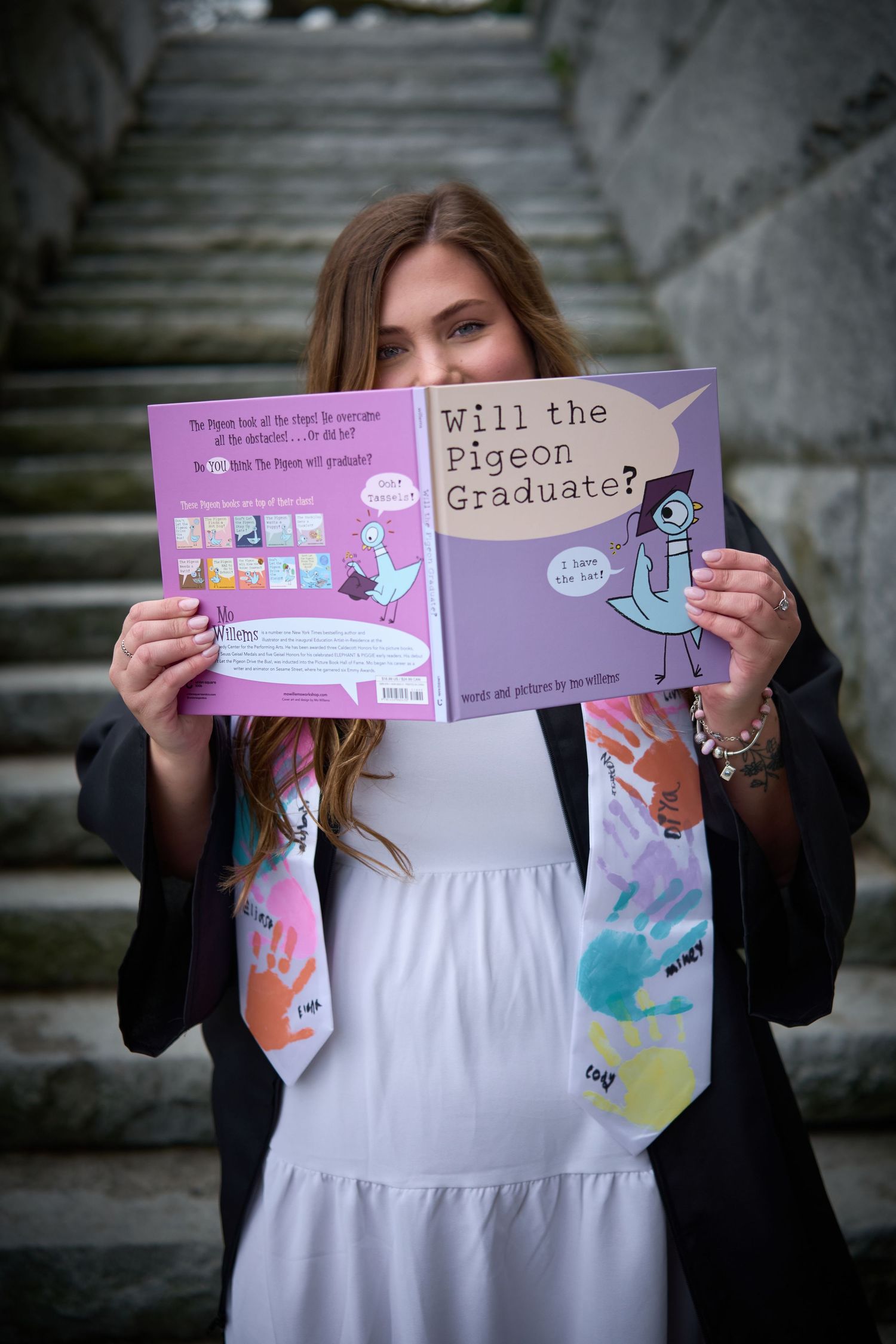 Graduate holding 'Will the Pigeon Graduate?' book, wearing cap and gown with colorful handprint stole on stone steps.