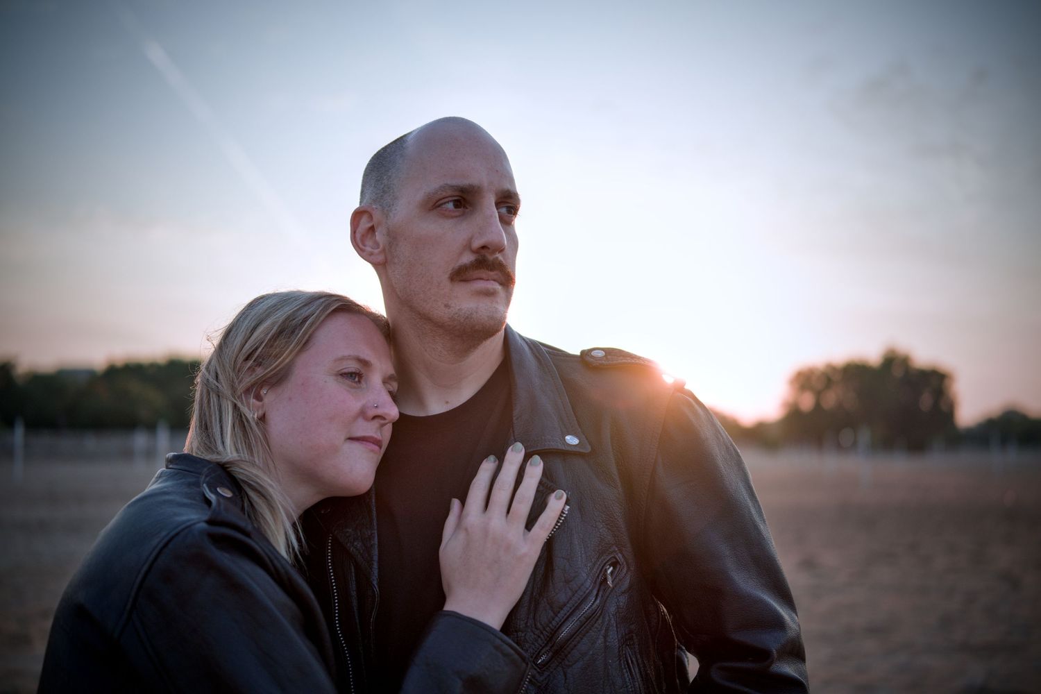 Couple in leather jackets sharing a tender moment outdoors at sunset, woman resting head on mans chest.