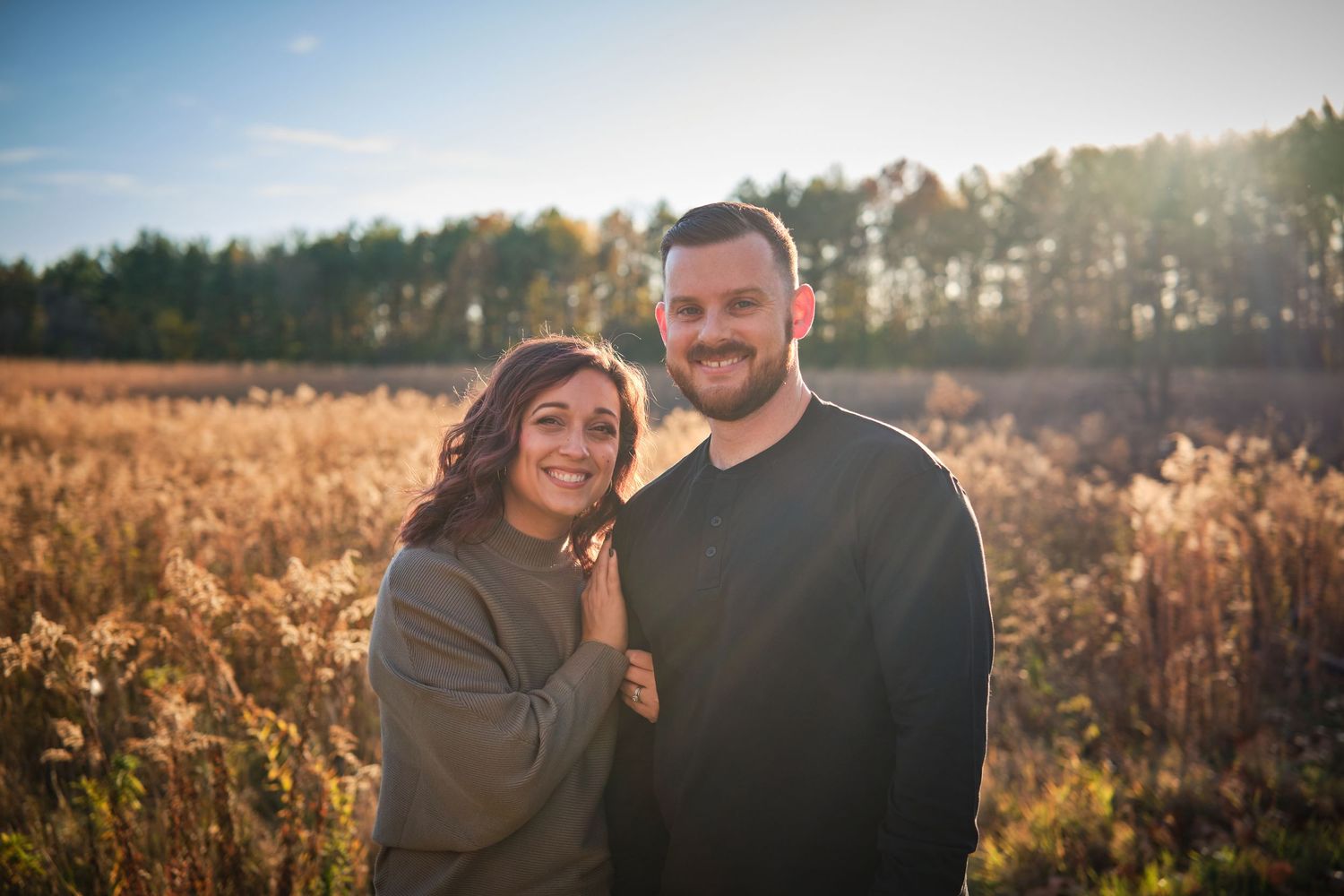 Couple smiling together in a sunlit autumn field with golden grasses and trees in the background.