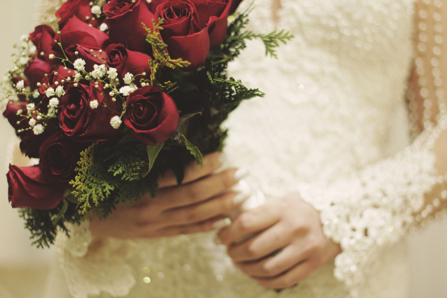 Close-up of hands holding a romantic red rose and baby's breath wedding bouquet against lace fabric.