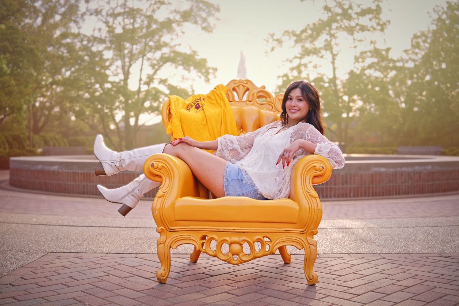 Young woman posing elegantly on a vibrant yellow ornate chair outdoors, wearing white boots and a lace top.