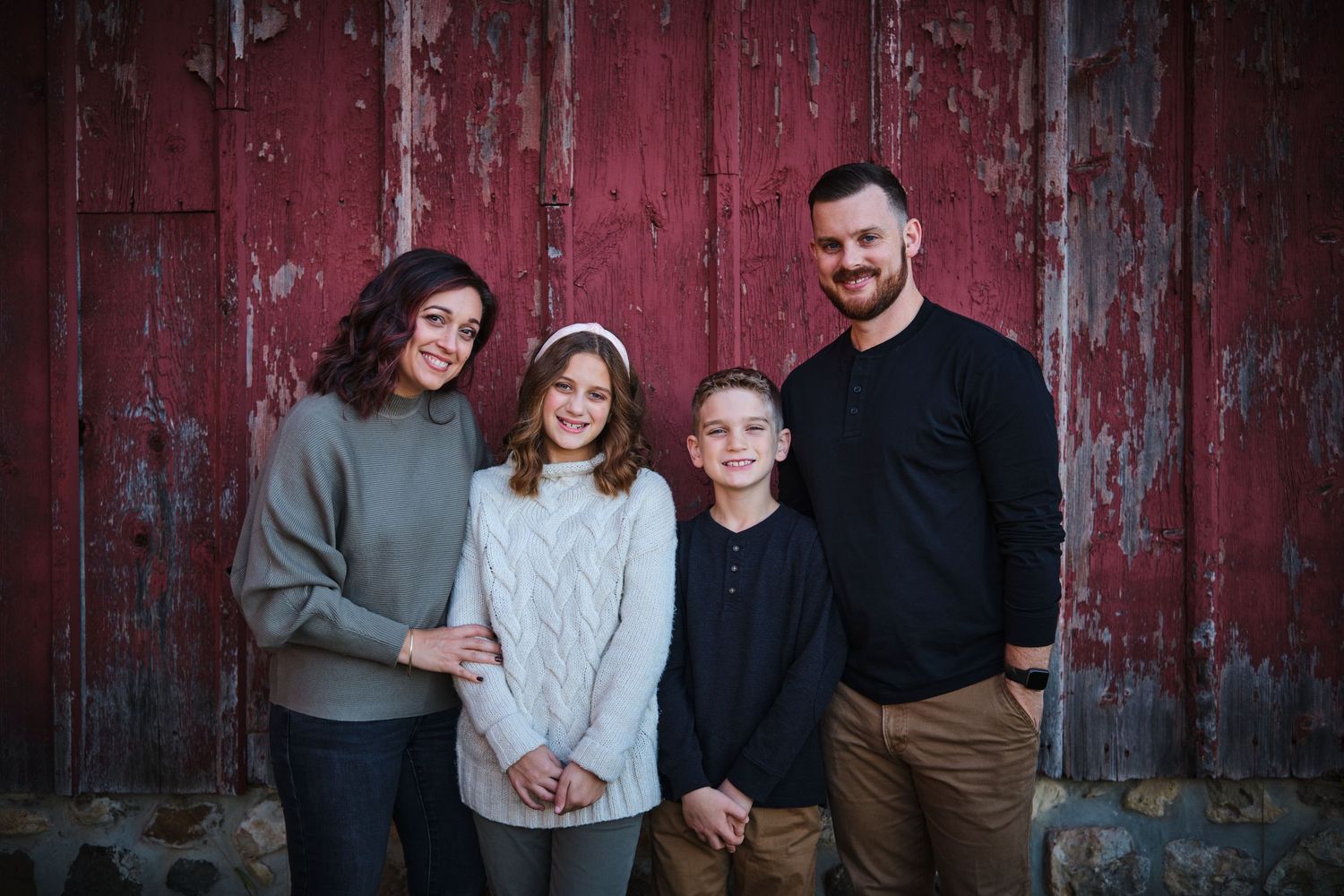 Family of four smiling together in front of a rustic red barn wall for a portrait photo.